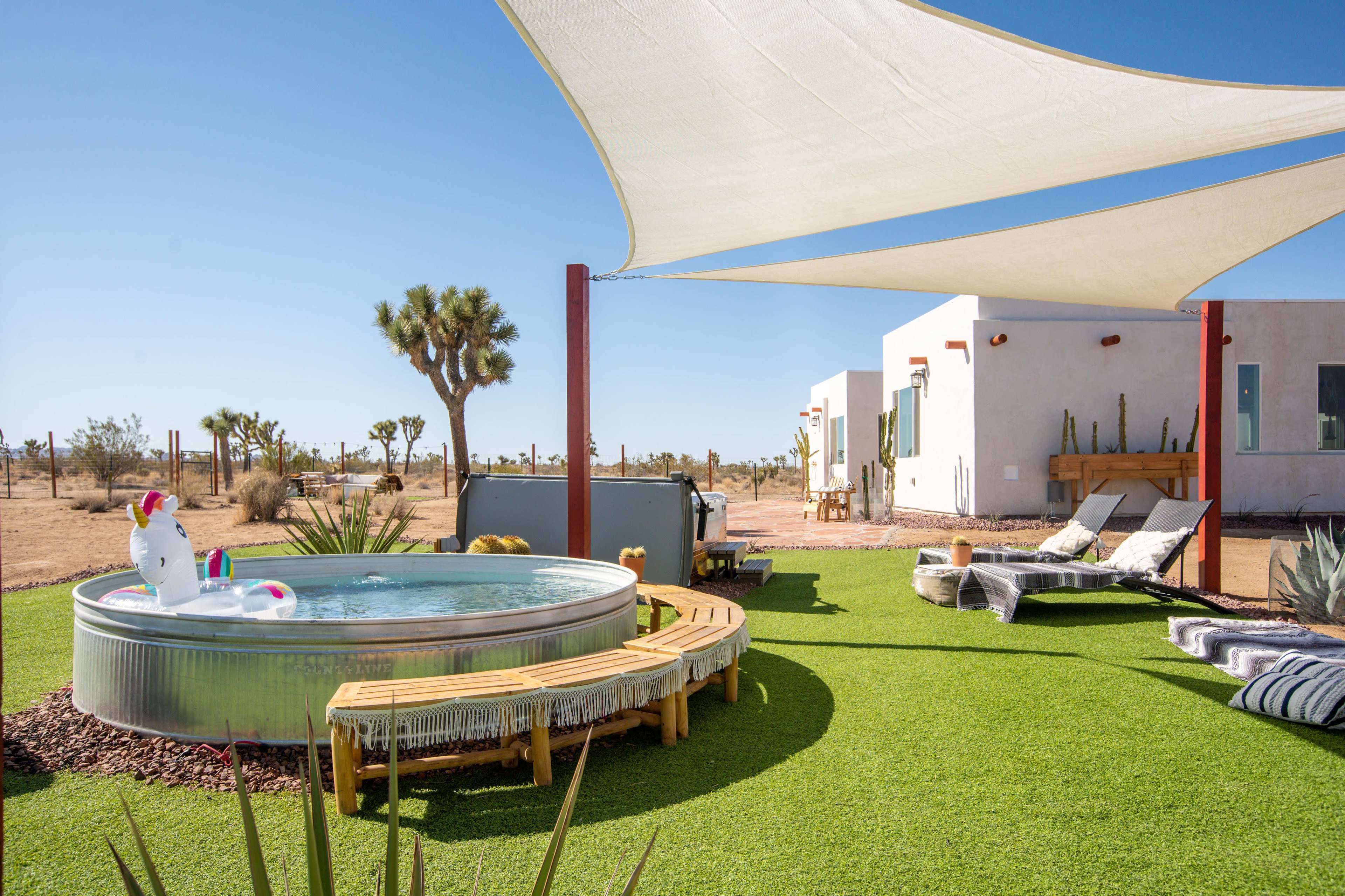 The image shows a small circular pool surrounded by a grassy area, with a shaded sitting space and desert landscape in the background.