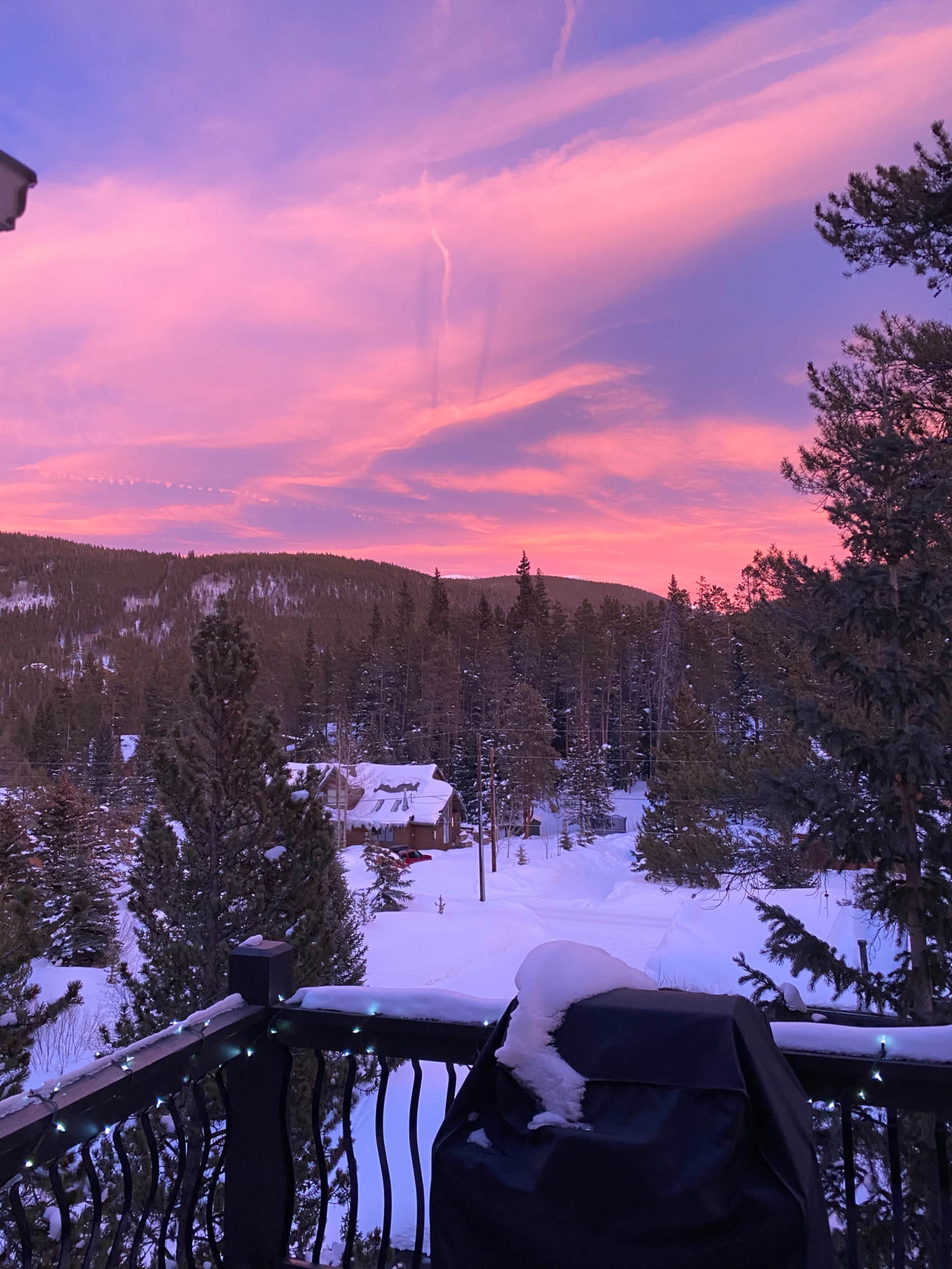A balcony view reveals a snow-covered landscape with a cabin surrounded by pine trees under a colorful sky at sunset.