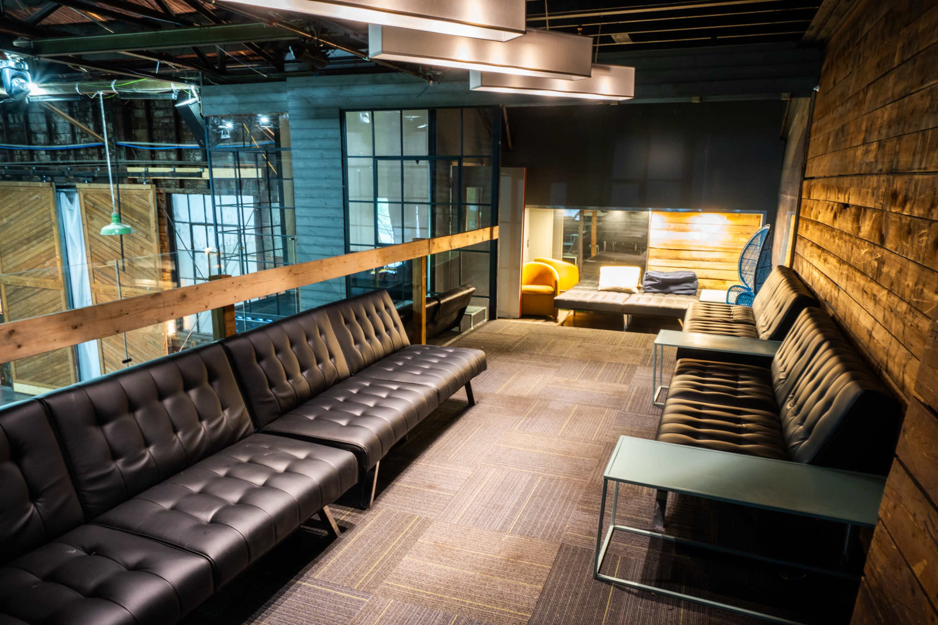 The image shows a modern lounge area with dark leather couches, wooden walls, and various seating options, illuminated by overhead lighting.