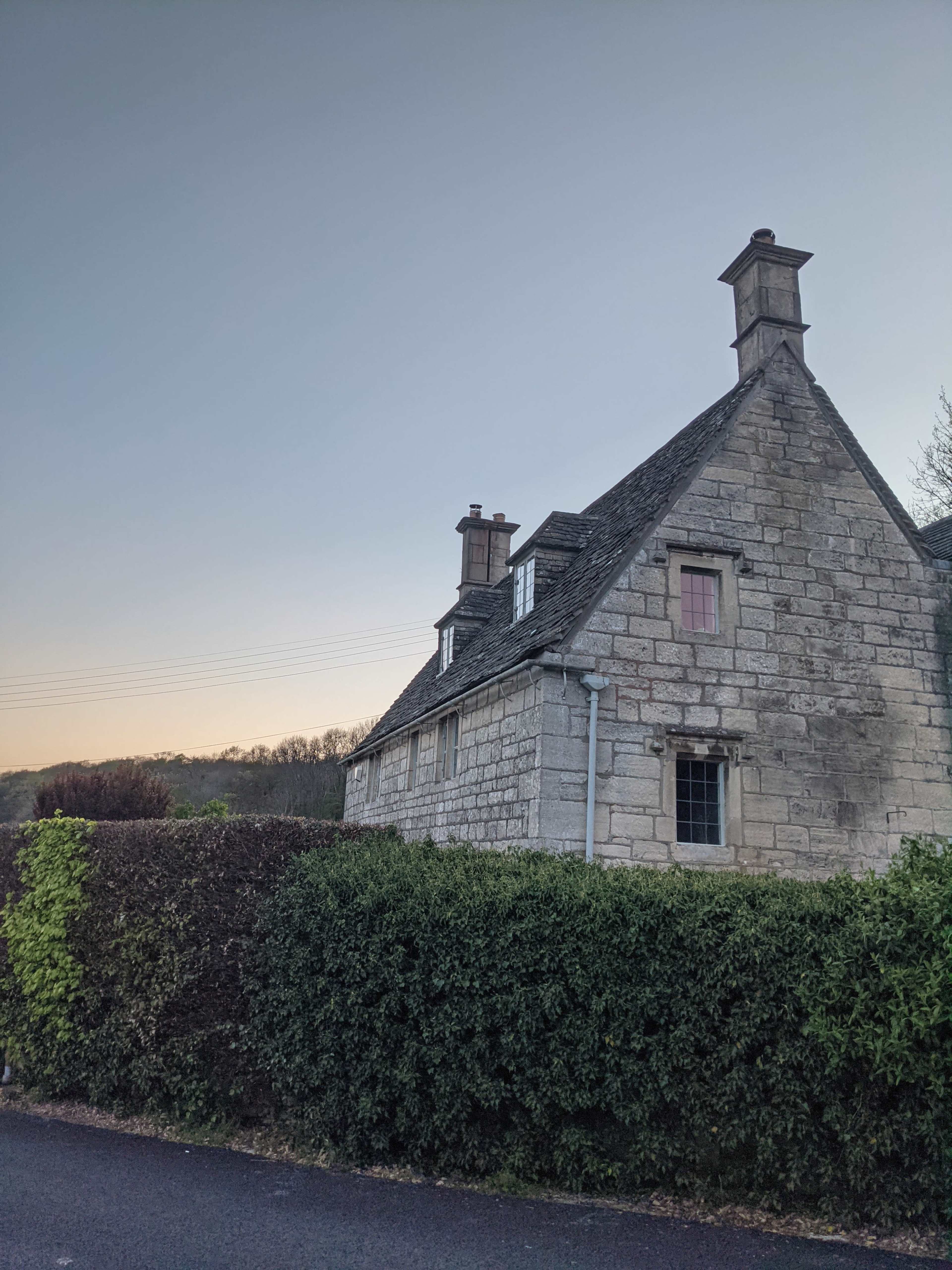 A stone house with a steep roof and chimneys is partially obscured by a hedge at dusk.