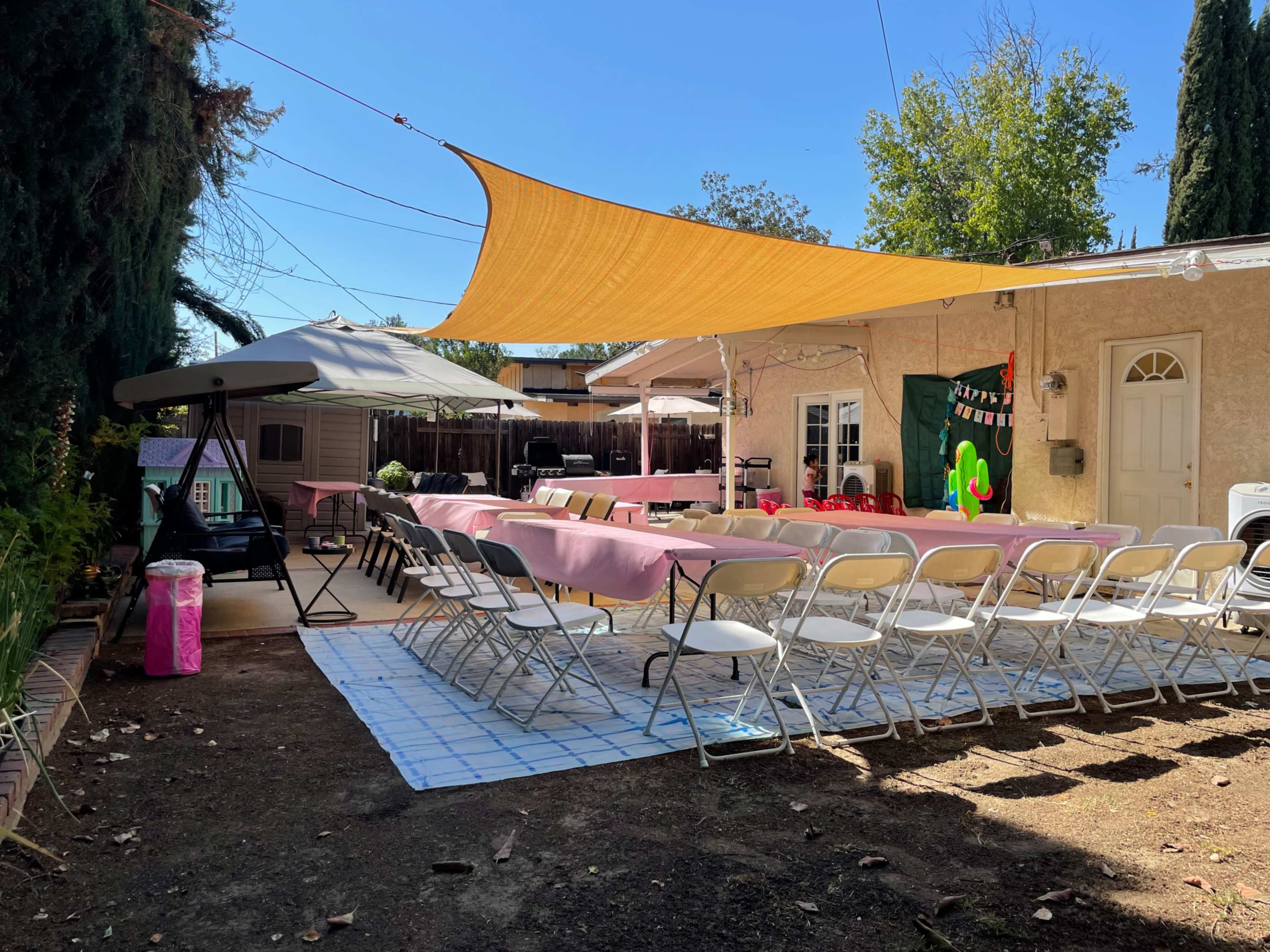 A backyard is set up for a gathering, featuring a long table covered with pink cloth, surrounded by white chairs, and shaded by a large orange canopy.