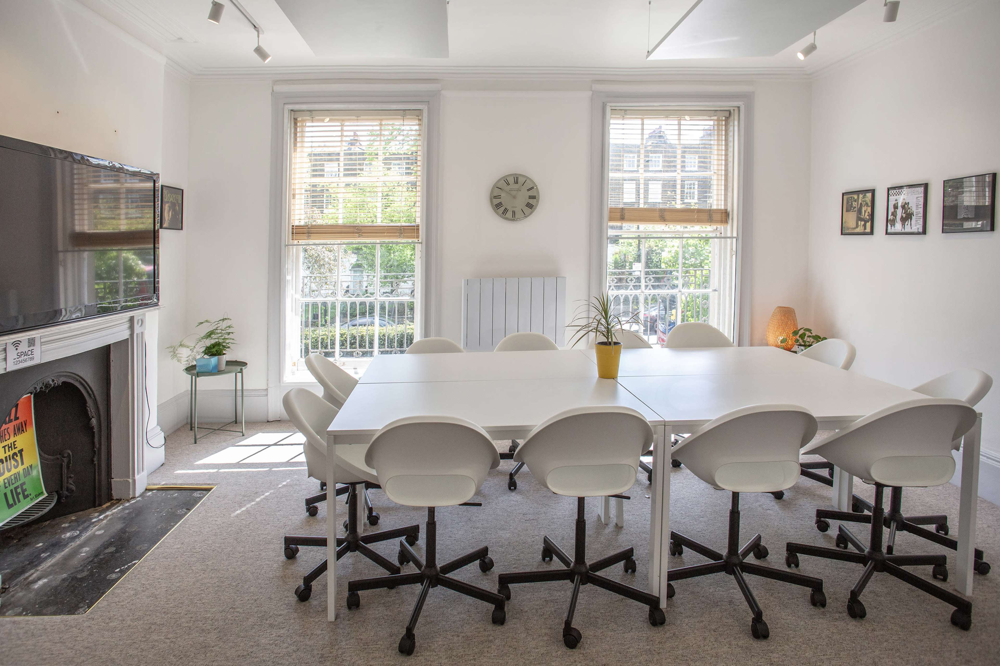 A bright meeting room features a large white table surrounded by chairs, with windows overlooking greenery and a clock on the wall.