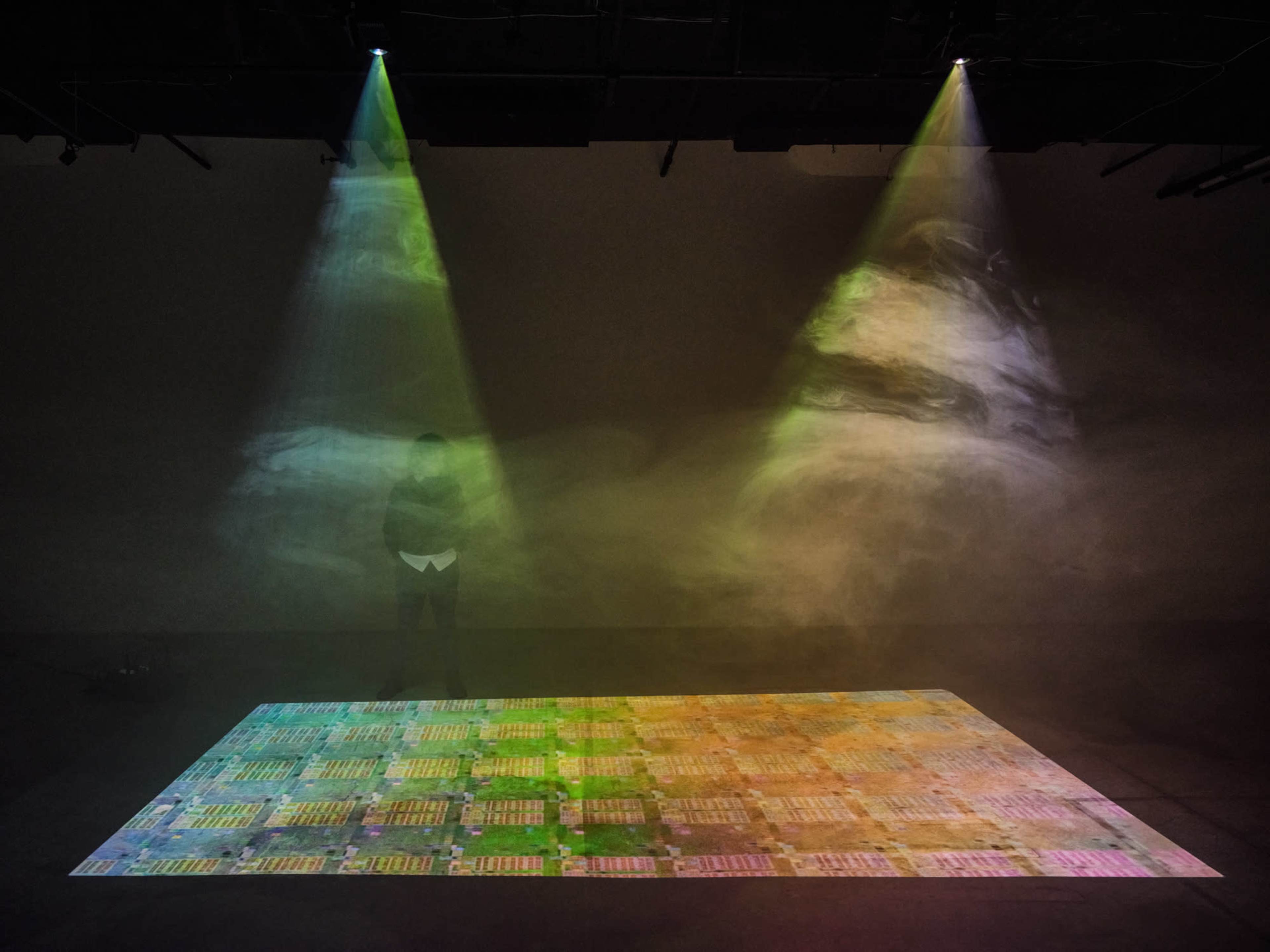 A person stands on a projection that displays a colorful, patterned surface illuminated by beams of light from above.