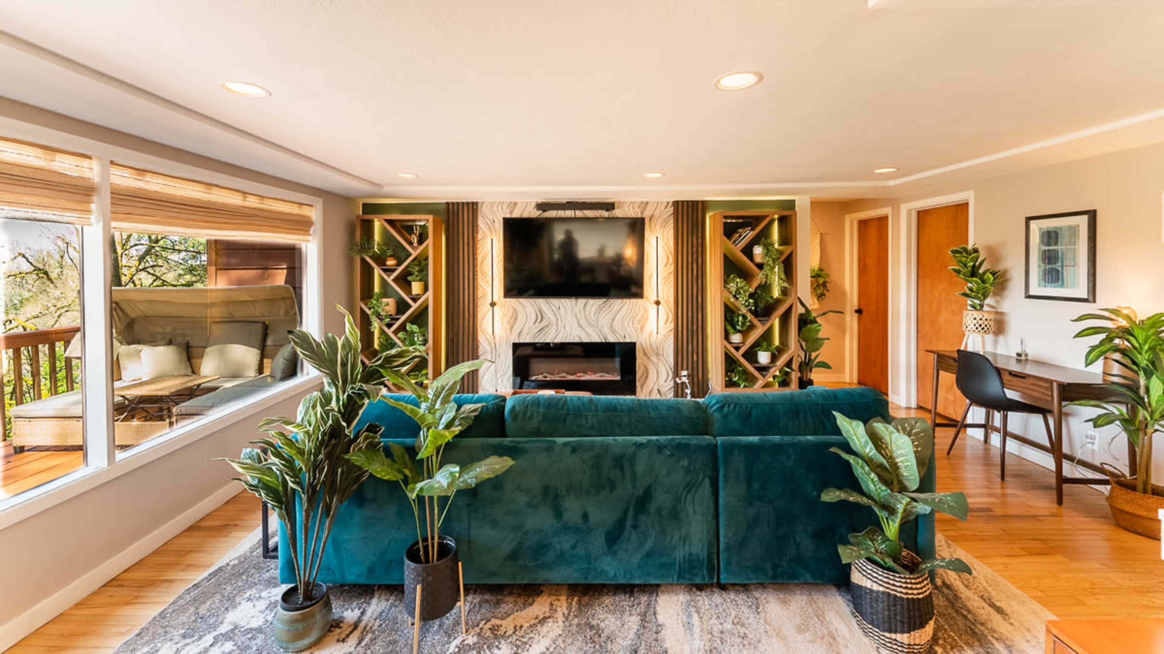 The living room features a teal sectional sofa facing a wall-mounted TV, surrounded by plants and bookshelves, with large windows letting in natural light.