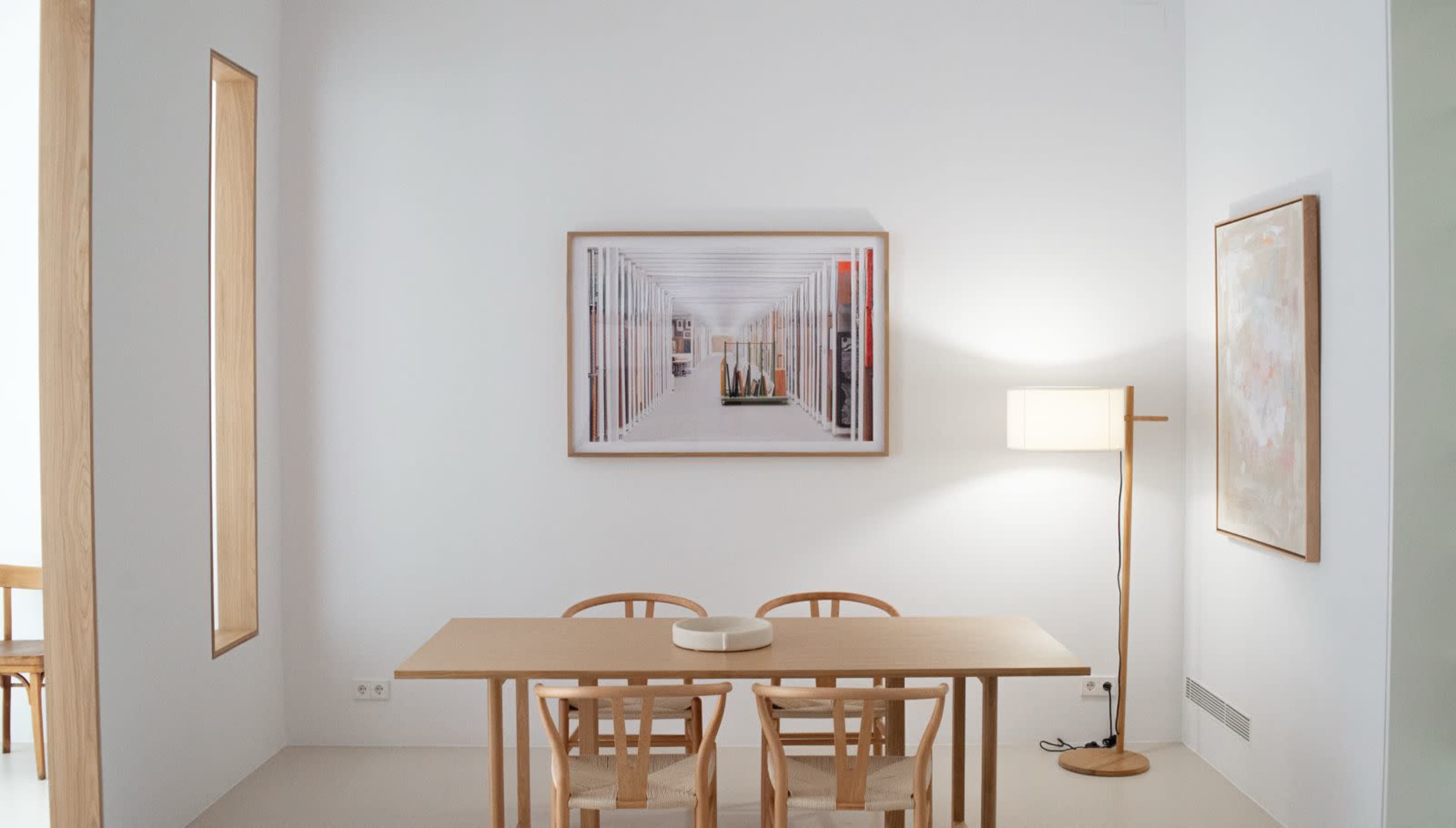 The image shows a minimalistic dining area featuring a wooden table and three chairs, with framed artwork and a floor lamp against a white wall.