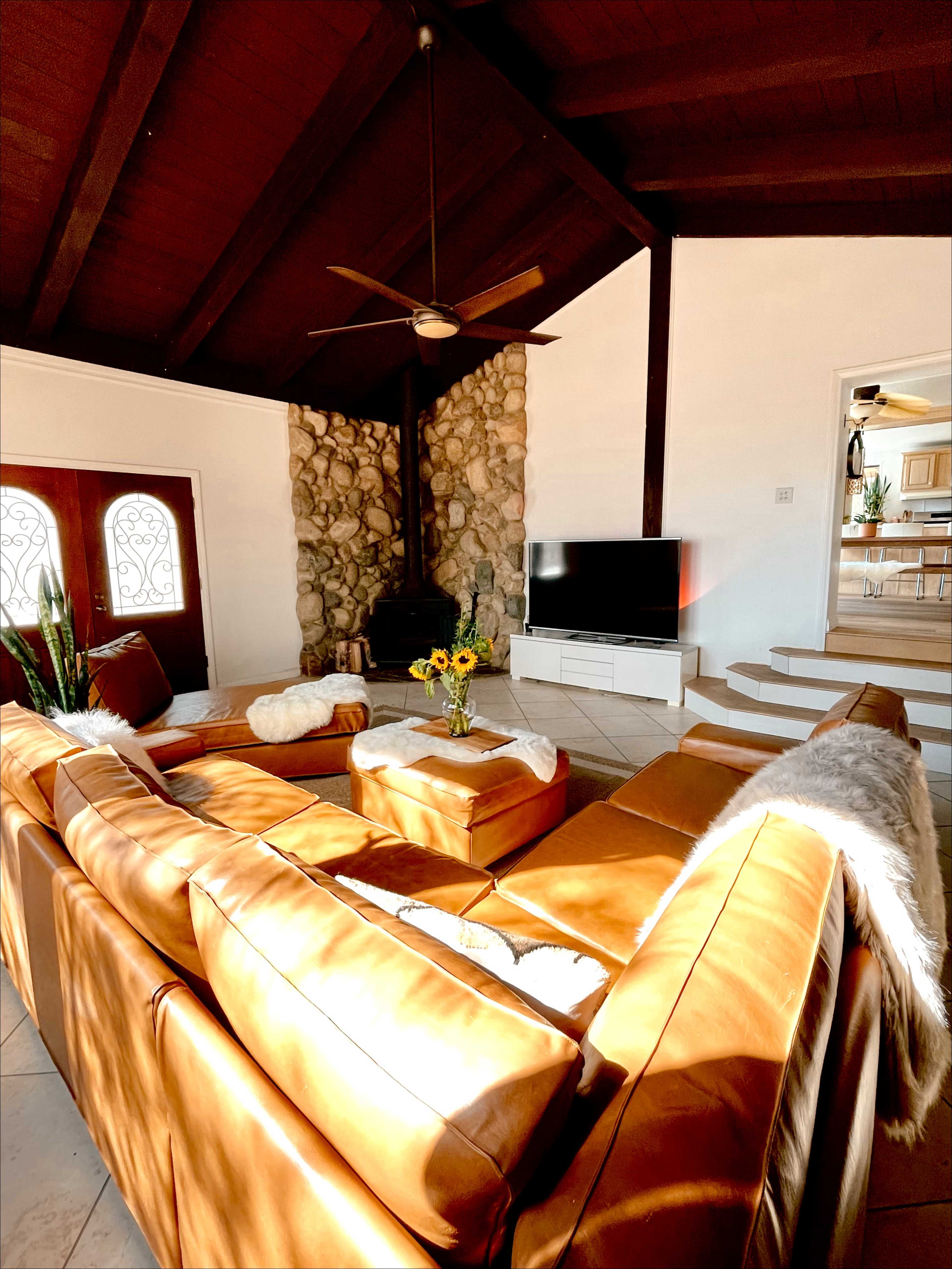 A spacious living room with a large brown leather sectional sofa, a coffee table with sunflowers, and a stone fireplace, all under a wood-beamed ceiling.