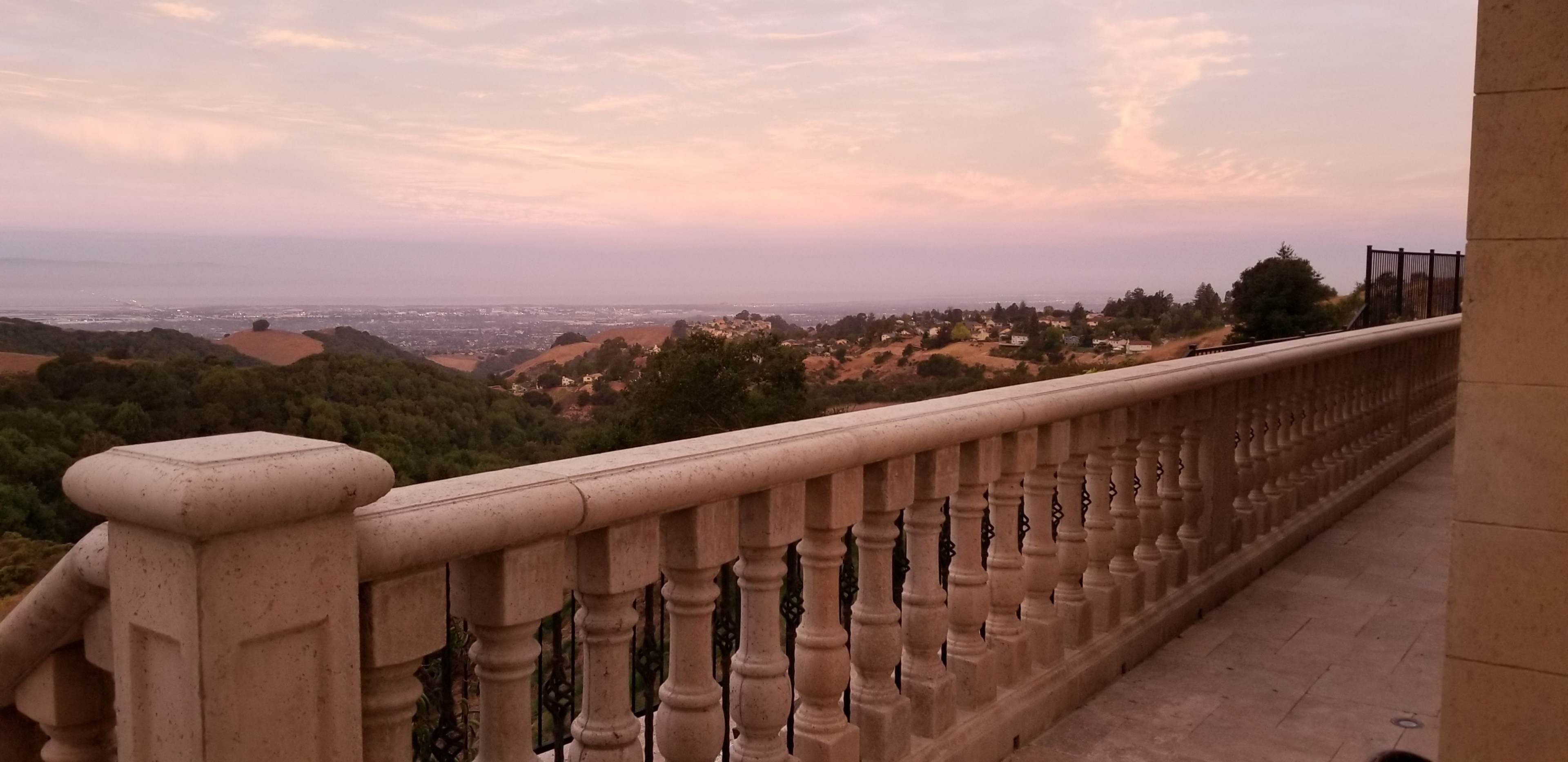 The image shows a stone balcony with ornate balusters overlooking a hilly landscape at dusk.