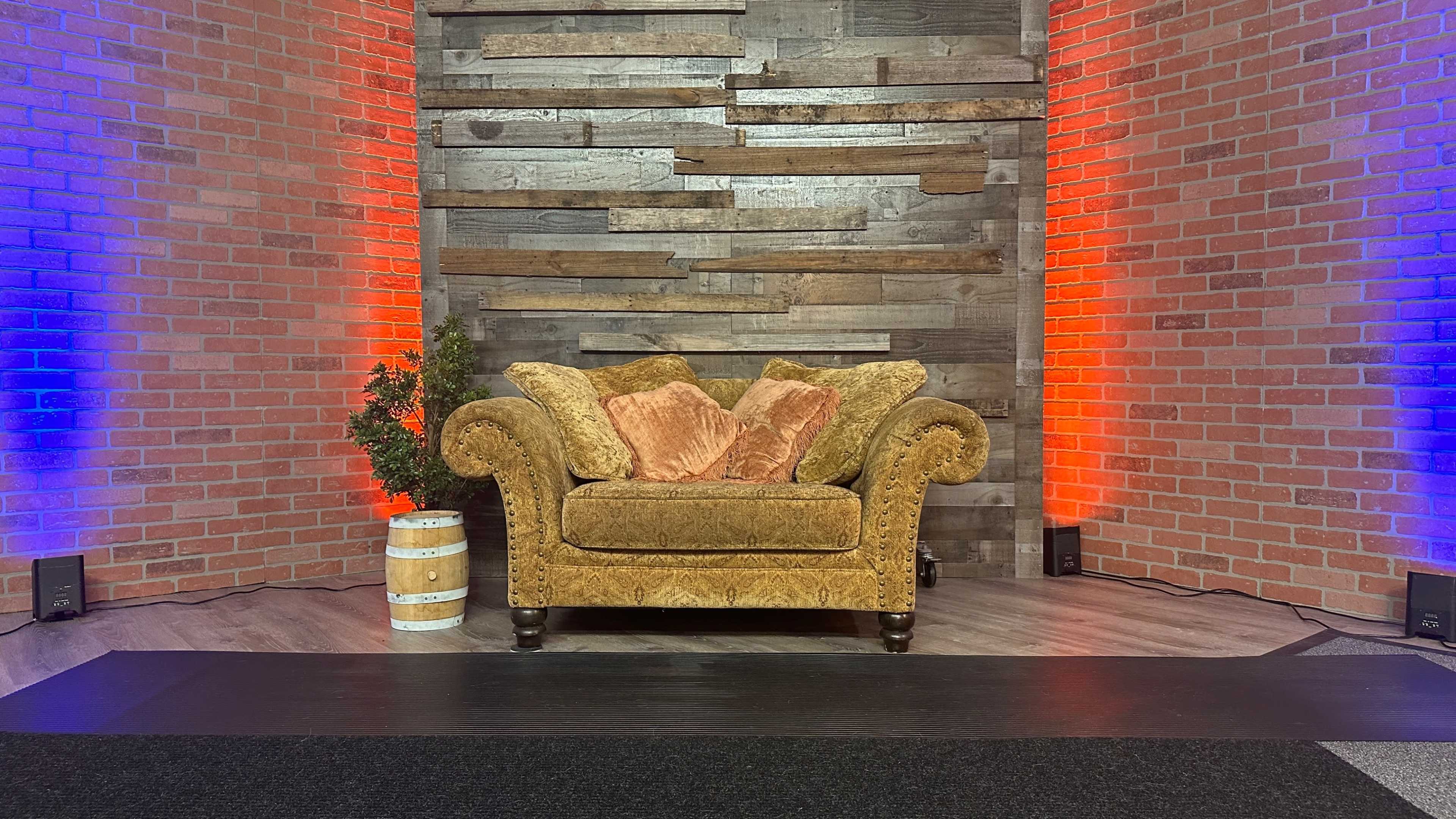 A brown, tufted couch with decorative throw pillows is positioned in front of a wooden wall and surrounded by red and blue lights.