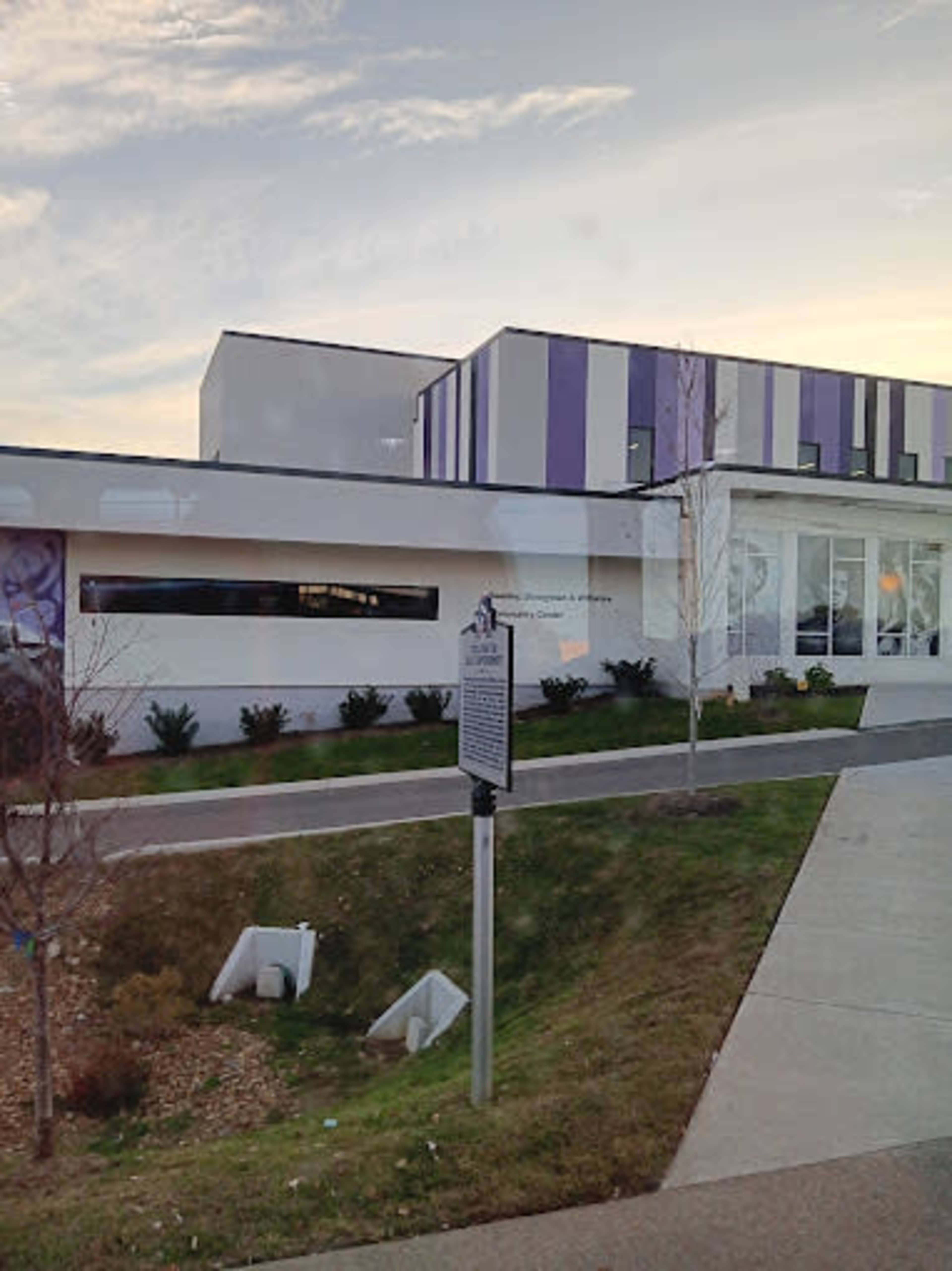 The image shows a modern building with a white exterior and purple accents, situated alongside a grassy area and pathway.