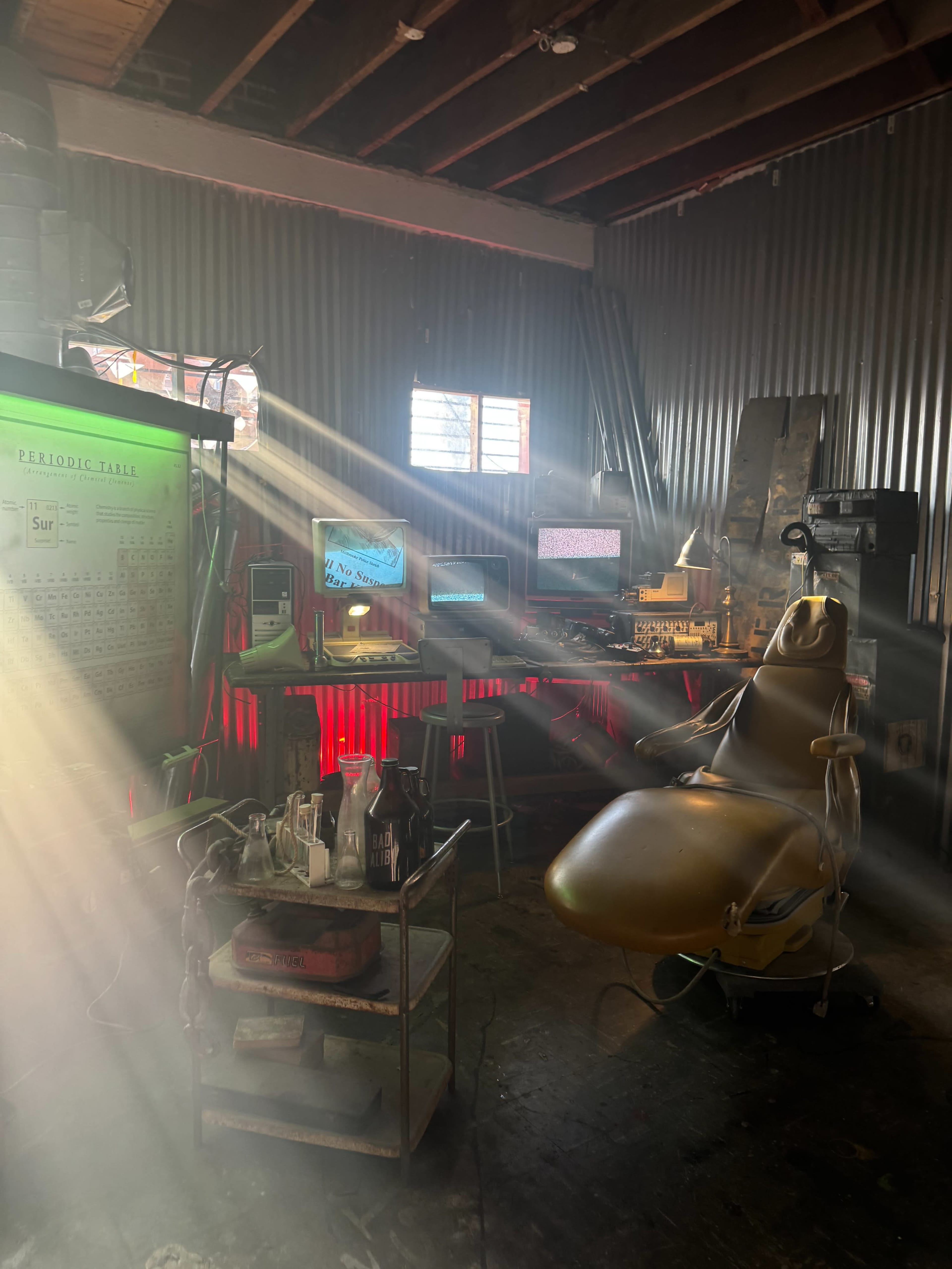 The image shows a dimly-lit room featuring a dental chair, multiple computer monitors, and various equipment arranged on a cart, with beams of light streaming through a window.