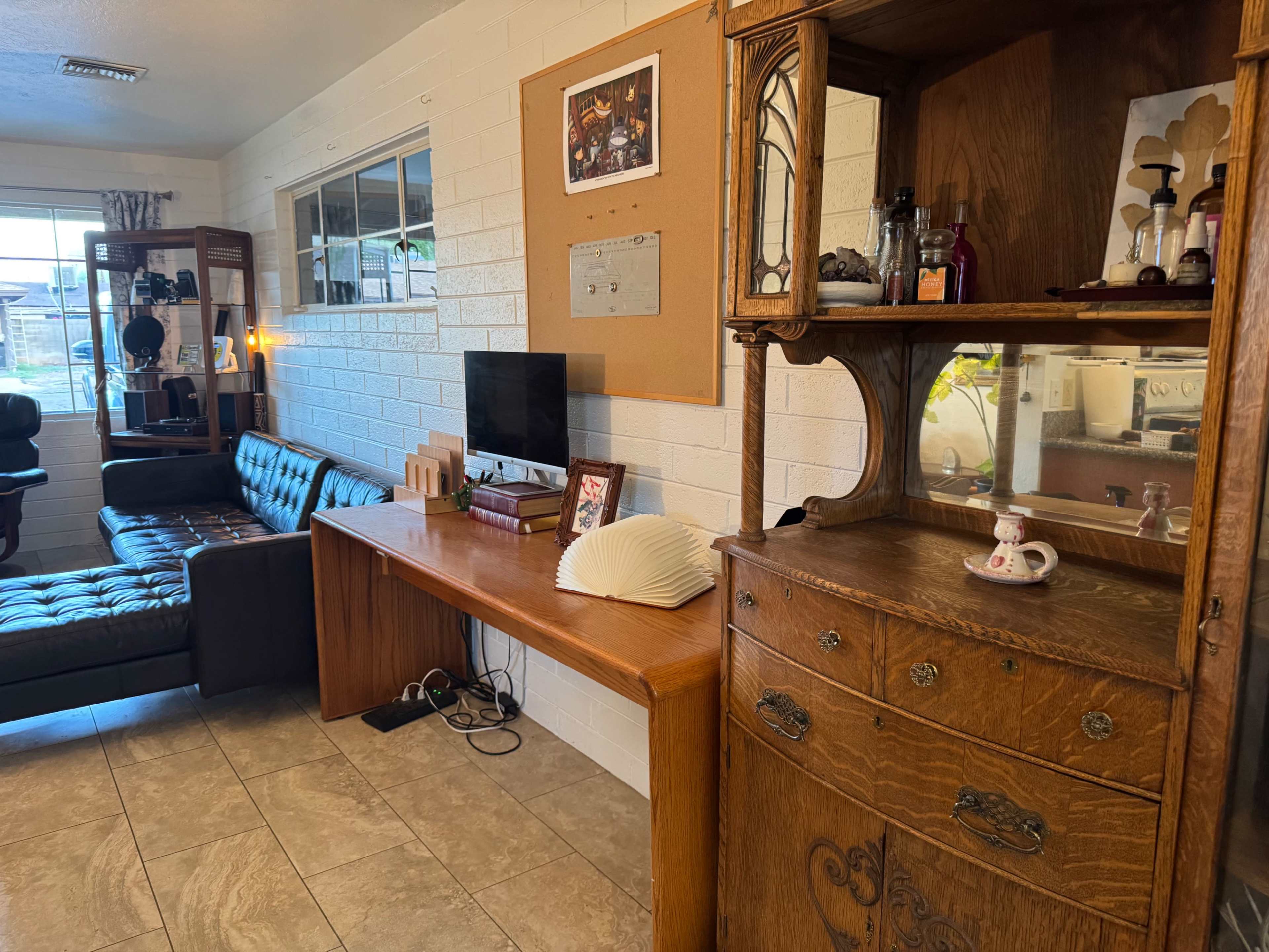 The image shows a living room with a black leather sofa, a wooden desk with a television, and a vintage wooden cabinet displaying decorative items.