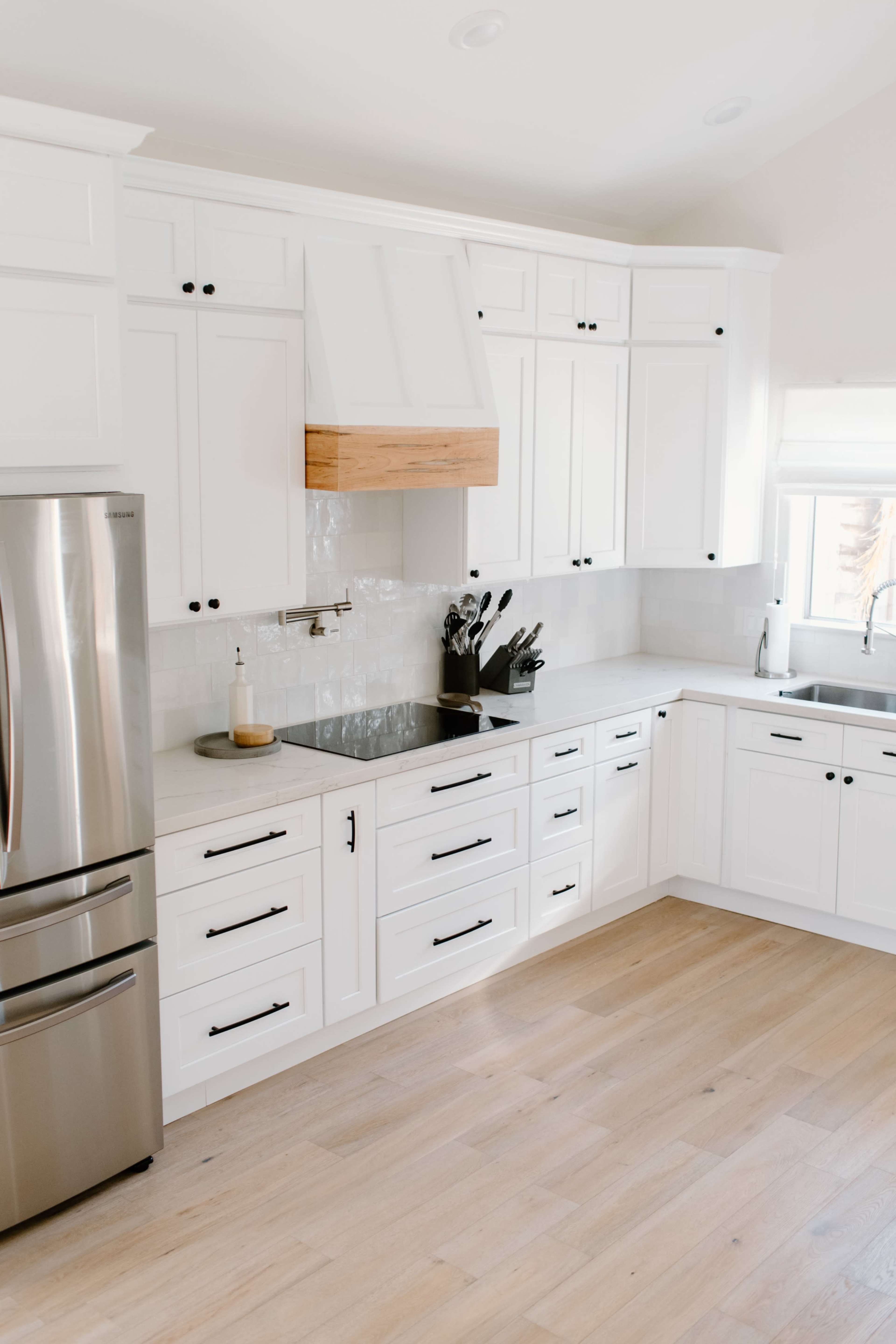 A modern kitchen features white cabinetry, a wooden hood over the stove, a stainless steel refrigerator, and a sink with natural light streaming in.