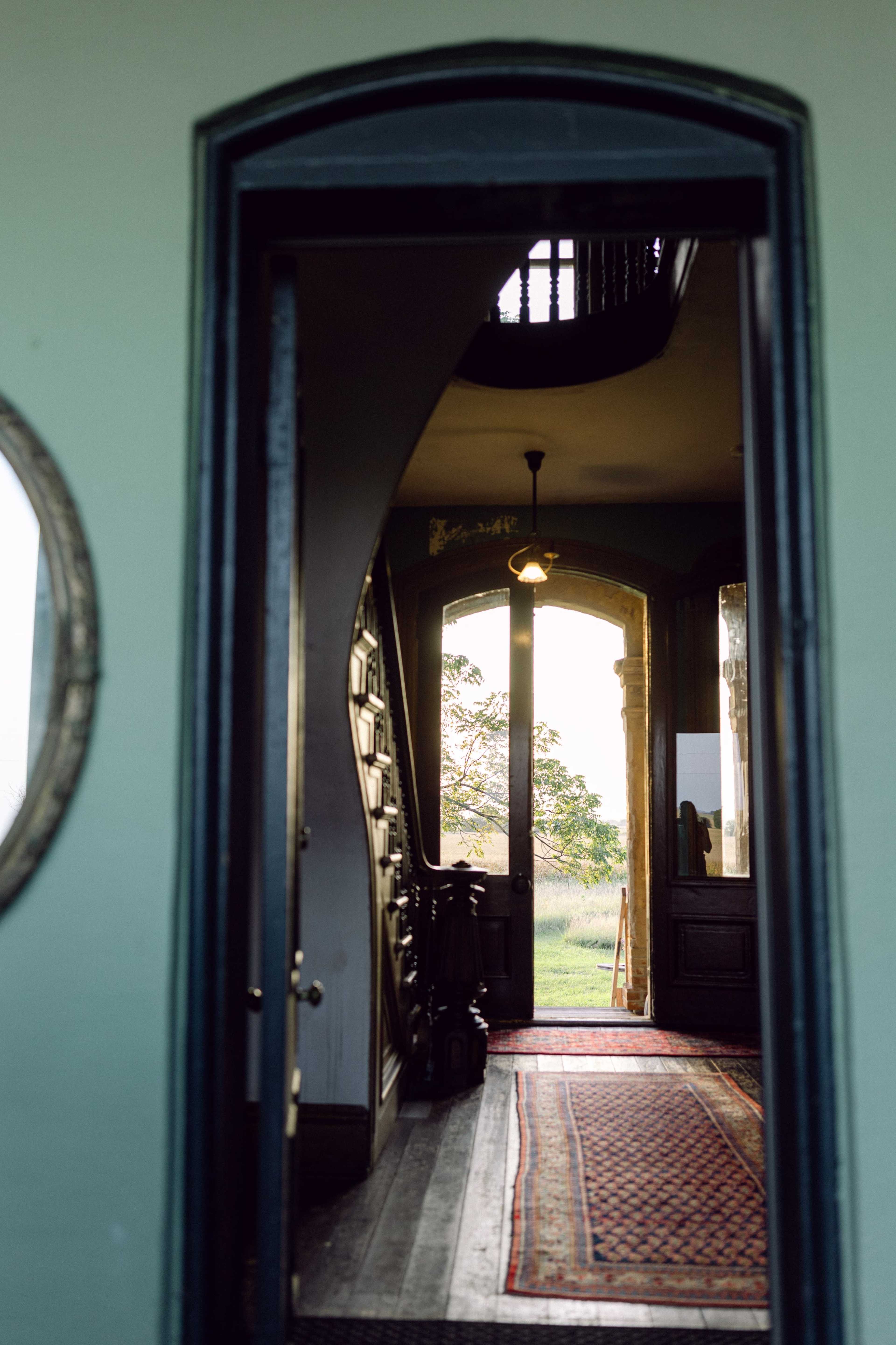 The image shows an entrance hallway with a door leading to an outdoor view, framed by a carpet and a decorative mirror on the wall.