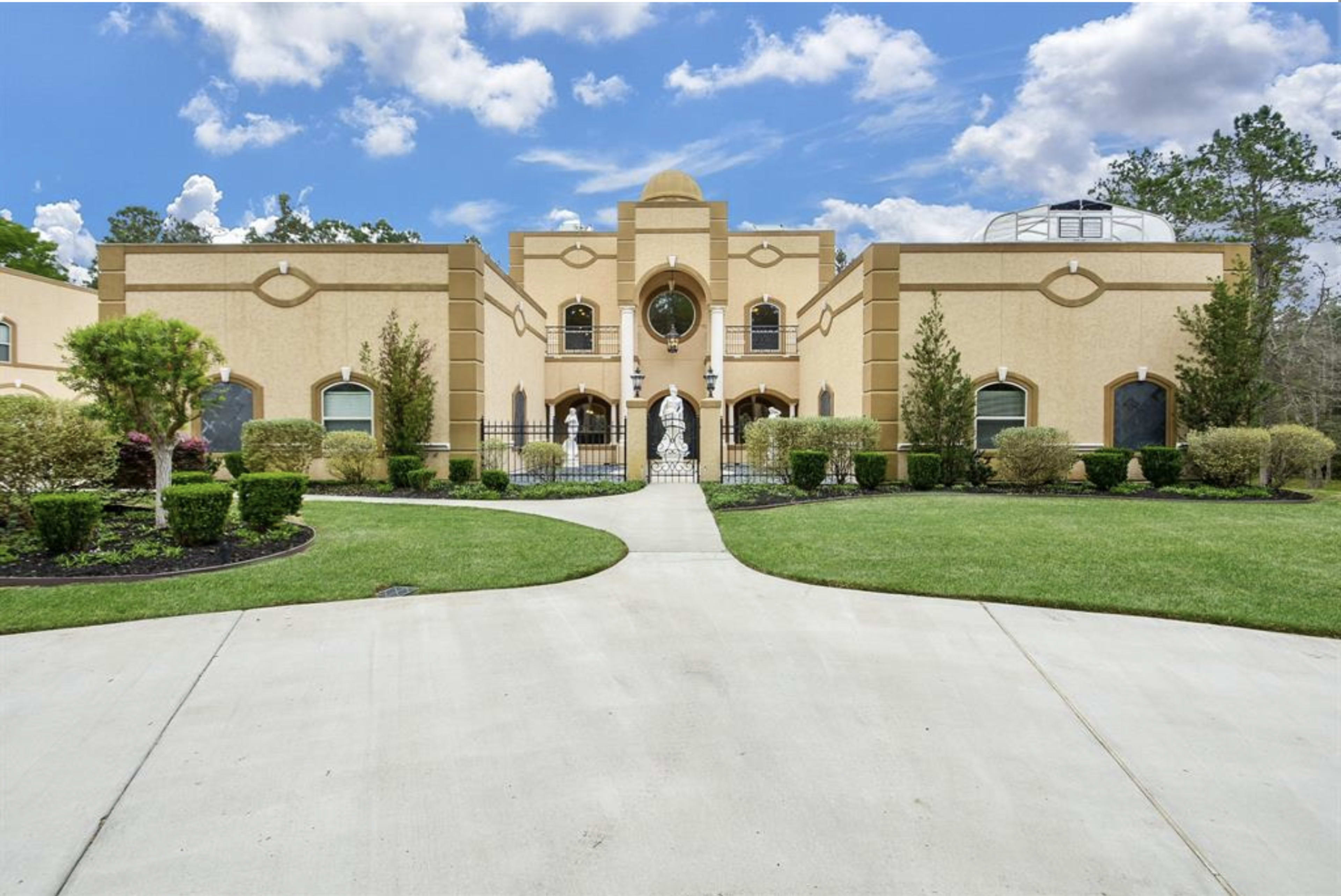 A large, ornate house framed by neatly trimmed shrubs and a paved pathway leading to a central entrance with a statue.