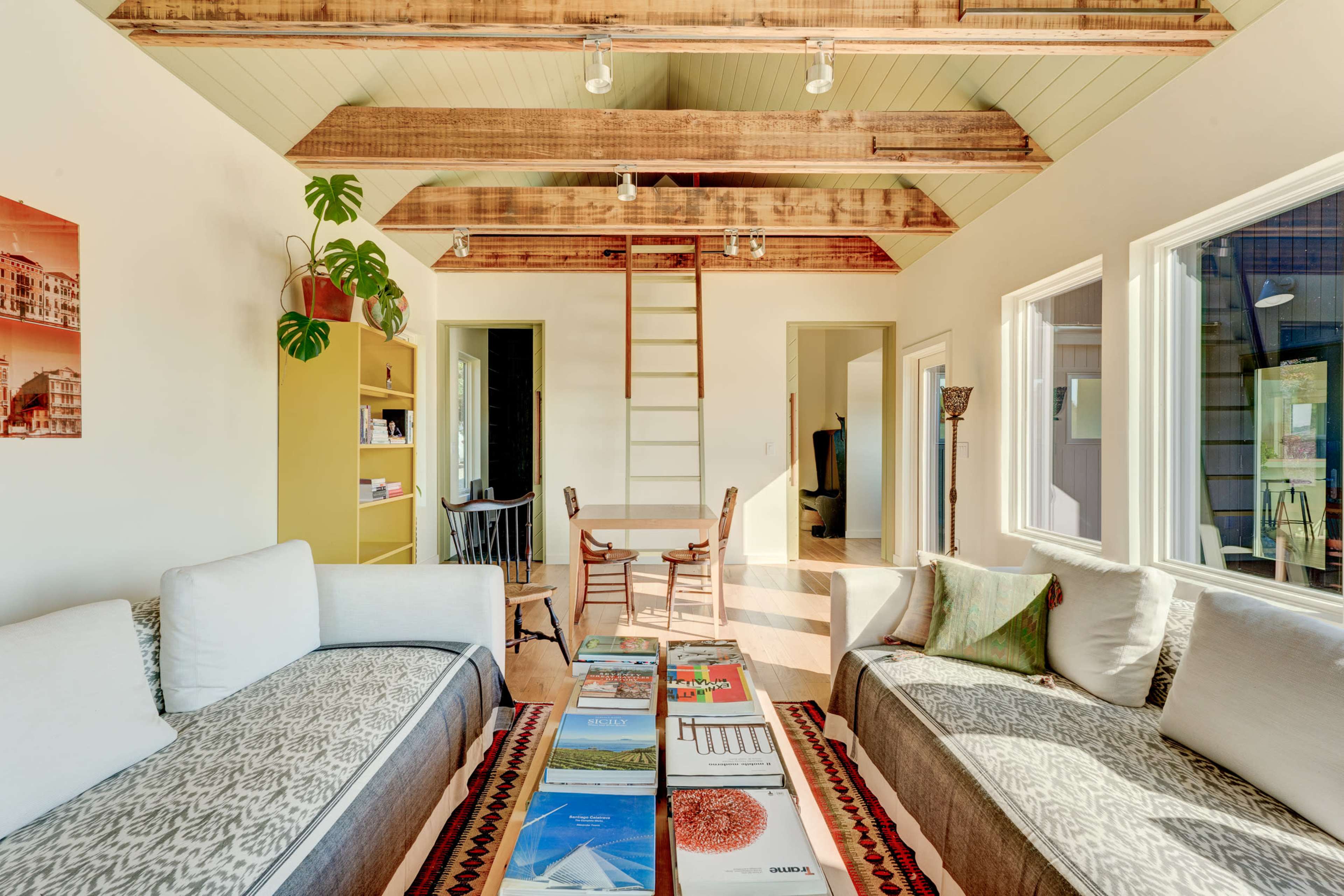 A modern living room features two sofas, a central coffee table with magazines, and a ladder leading to an upper level, surrounded by large windows.