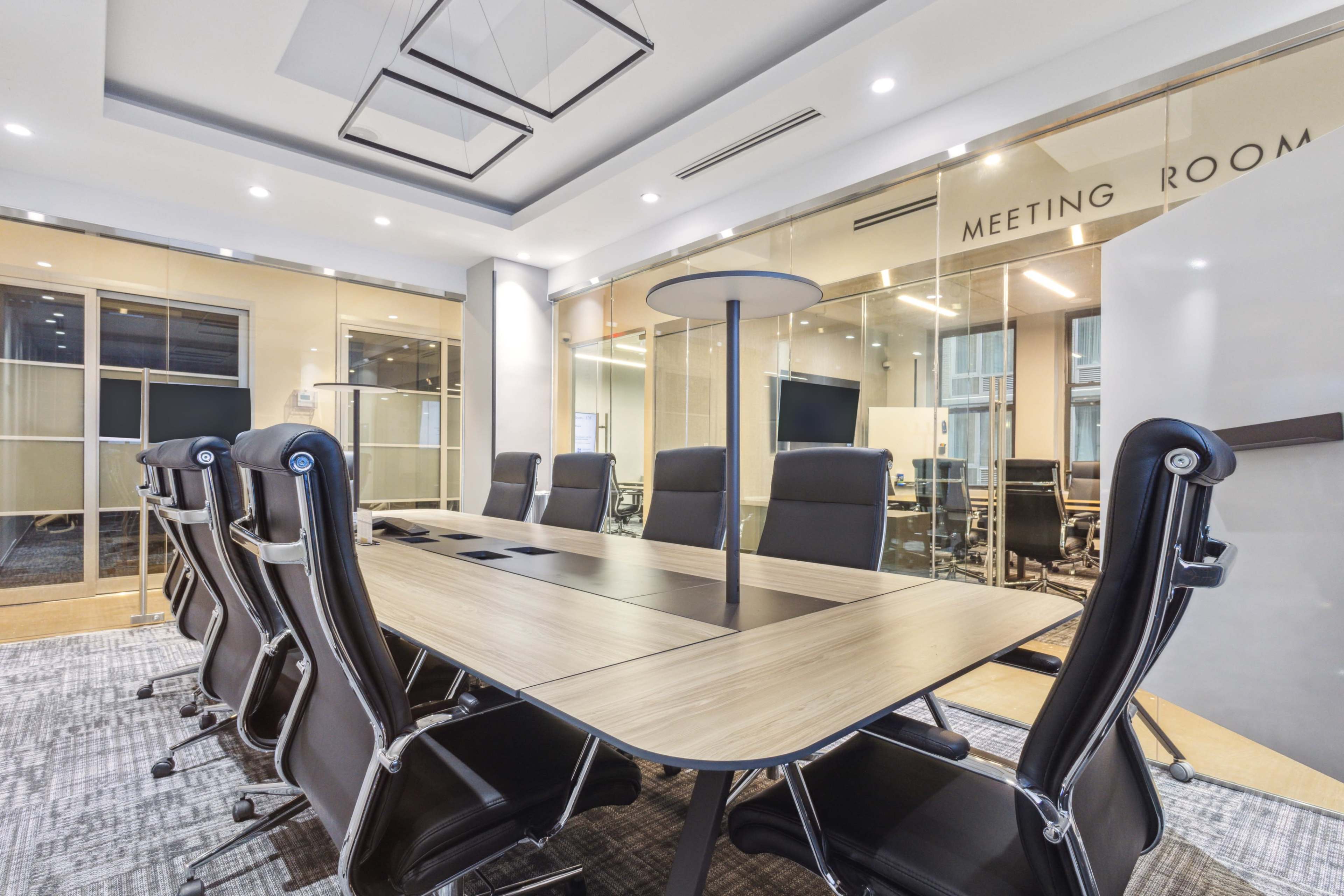 A modern conference room features a long wooden table surrounded by black ergonomic chairs, with glass walls and a large screen visible in the background.