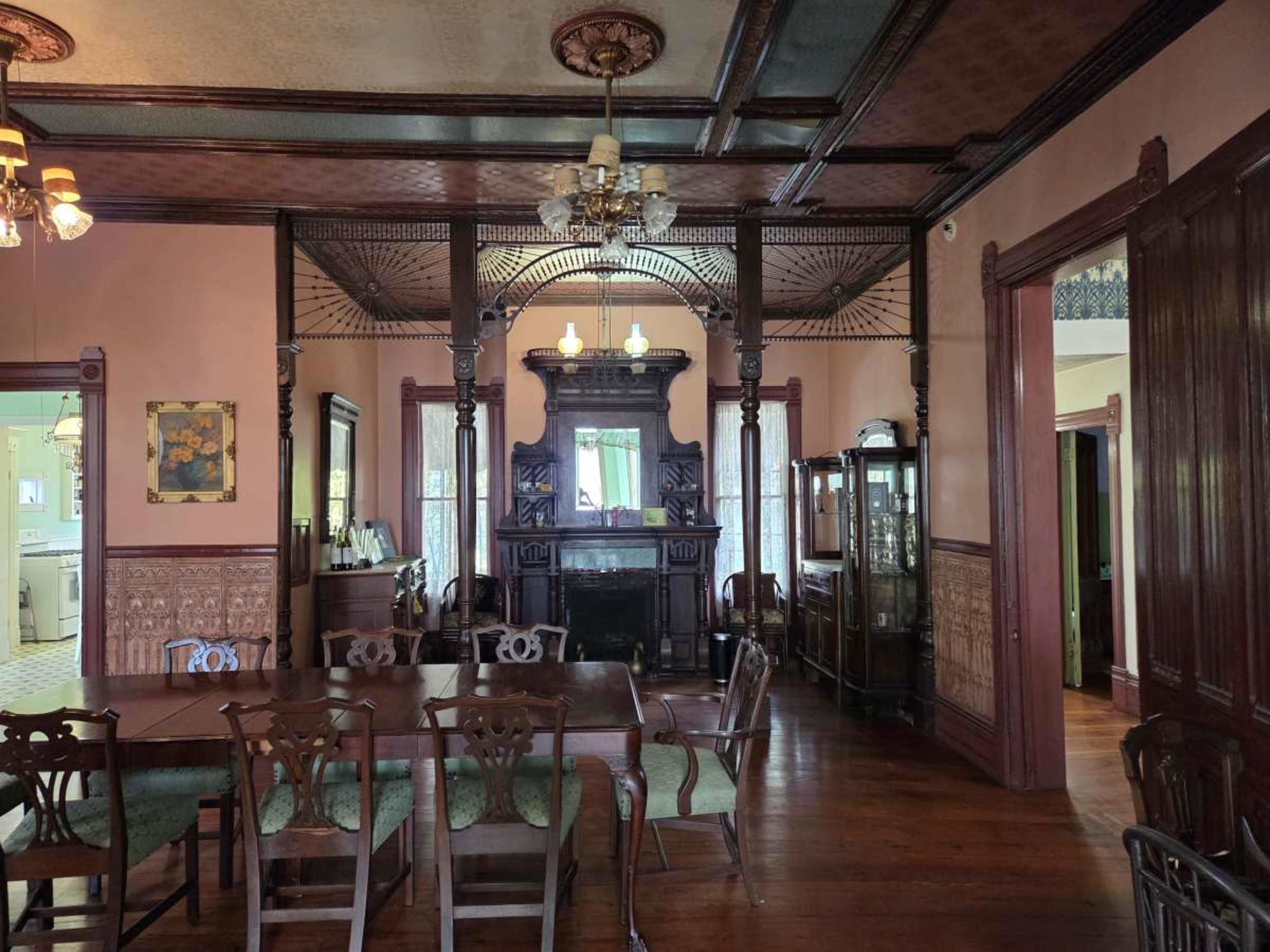 The image shows a vintage dining room with an intricate wooden dining table, chairs, and a large, ornate fireplace surrounded by decorative woodwork.