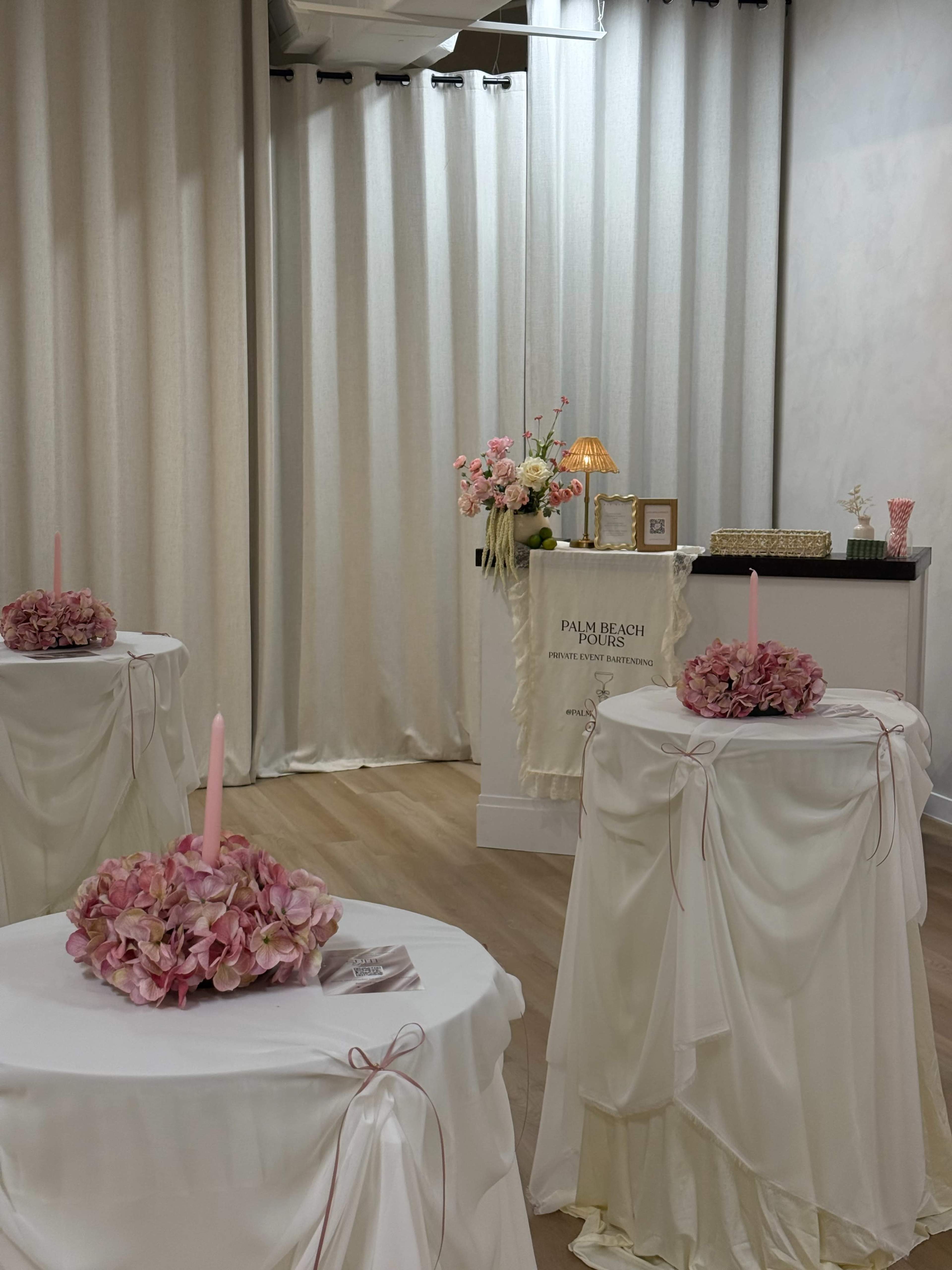 The image depicts an elegantly decorated interior featuring three tables with floral centerpieces and candles, alongside a reception area with a sign and floral arrangements.