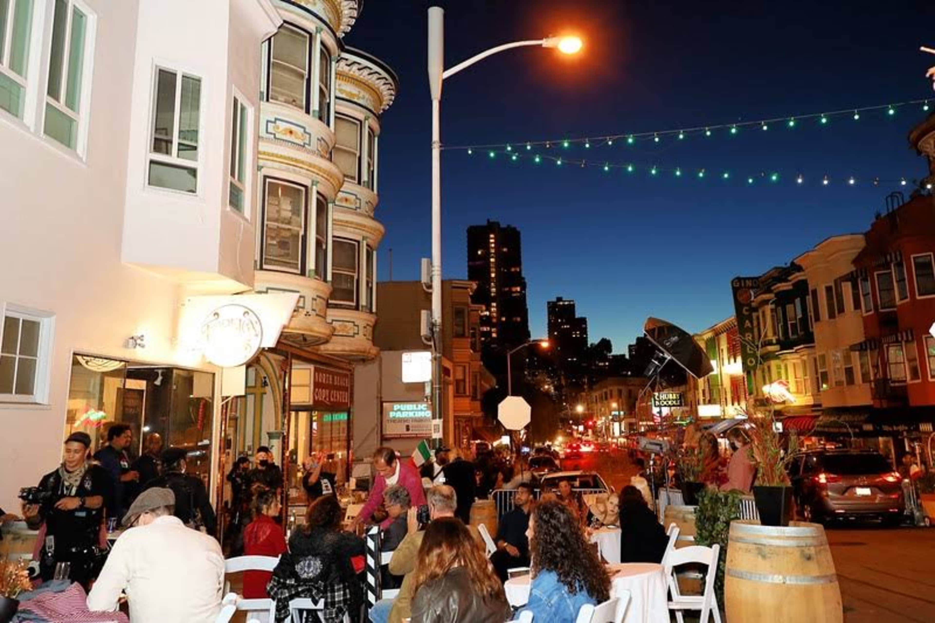The scene shows a lively outdoor dining area in an urban setting at dusk, with people seated at tables and city buildings illuminated in the background.