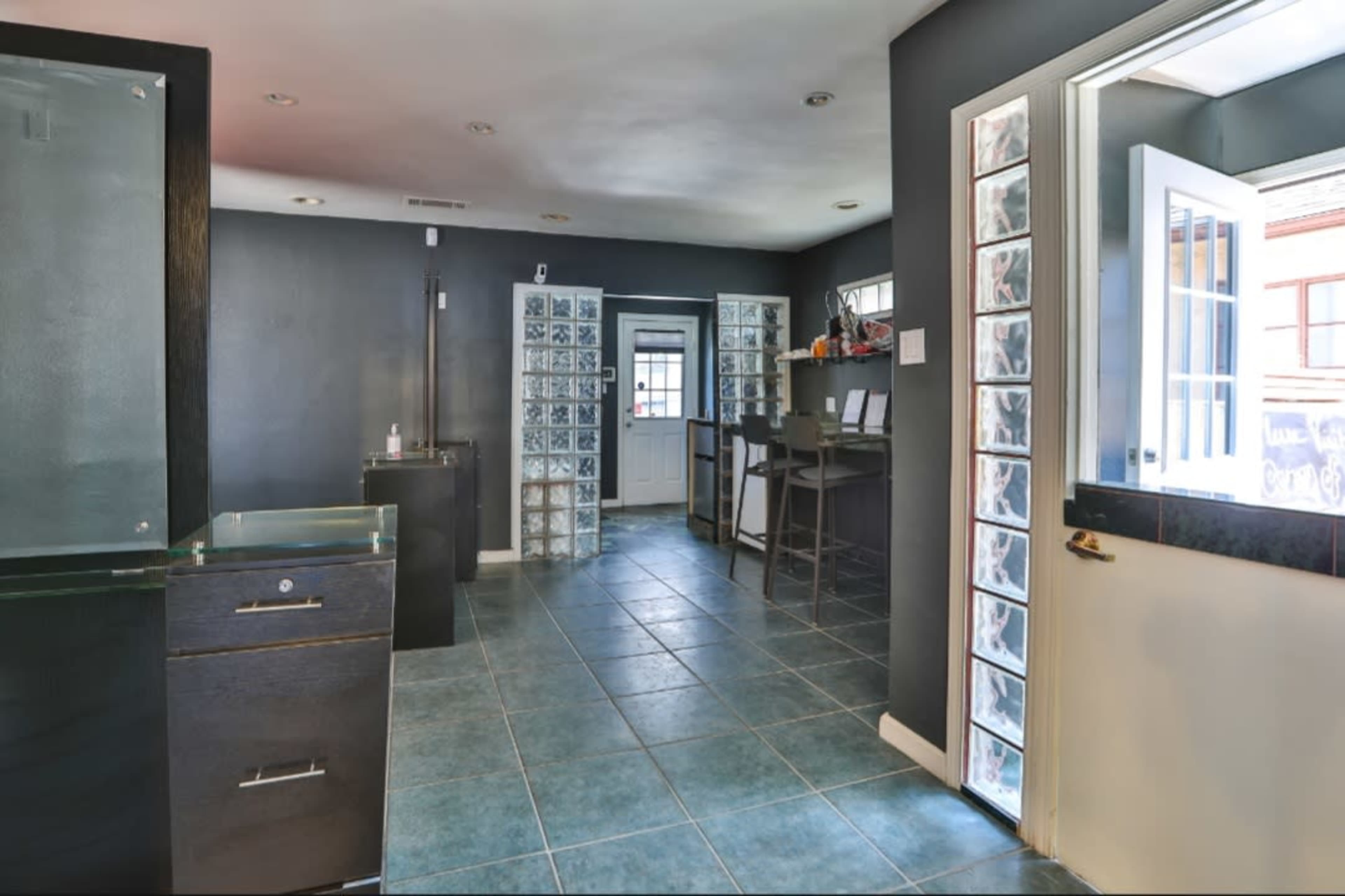 The image shows a spacious interior with dark walls and tiled flooring, featuring a doorway leading outside, glass block windows, and a bar-like counter.