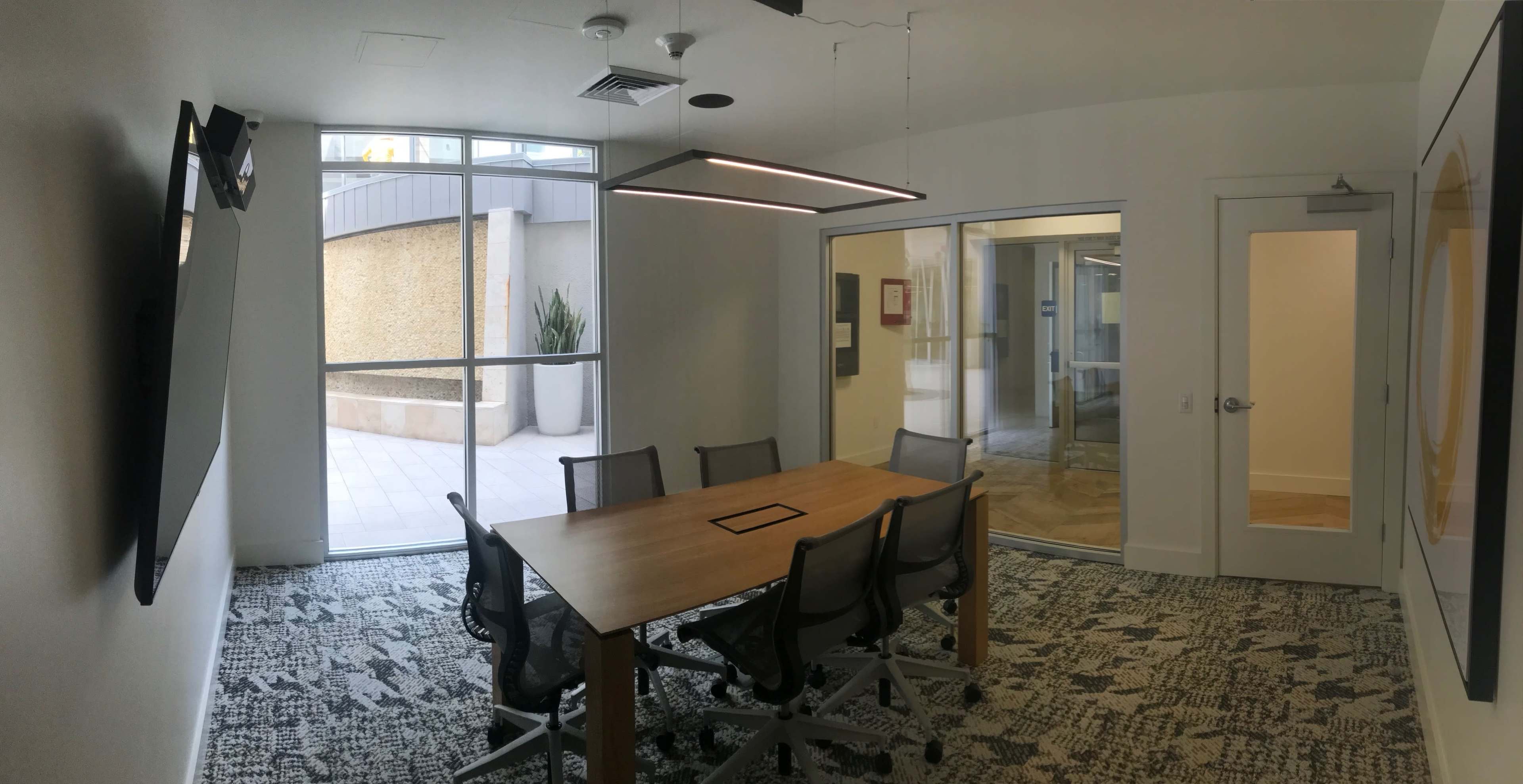 A modern conference room features a large wooden table surrounded by rolling chairs and glass walls that open to an outdoor area.