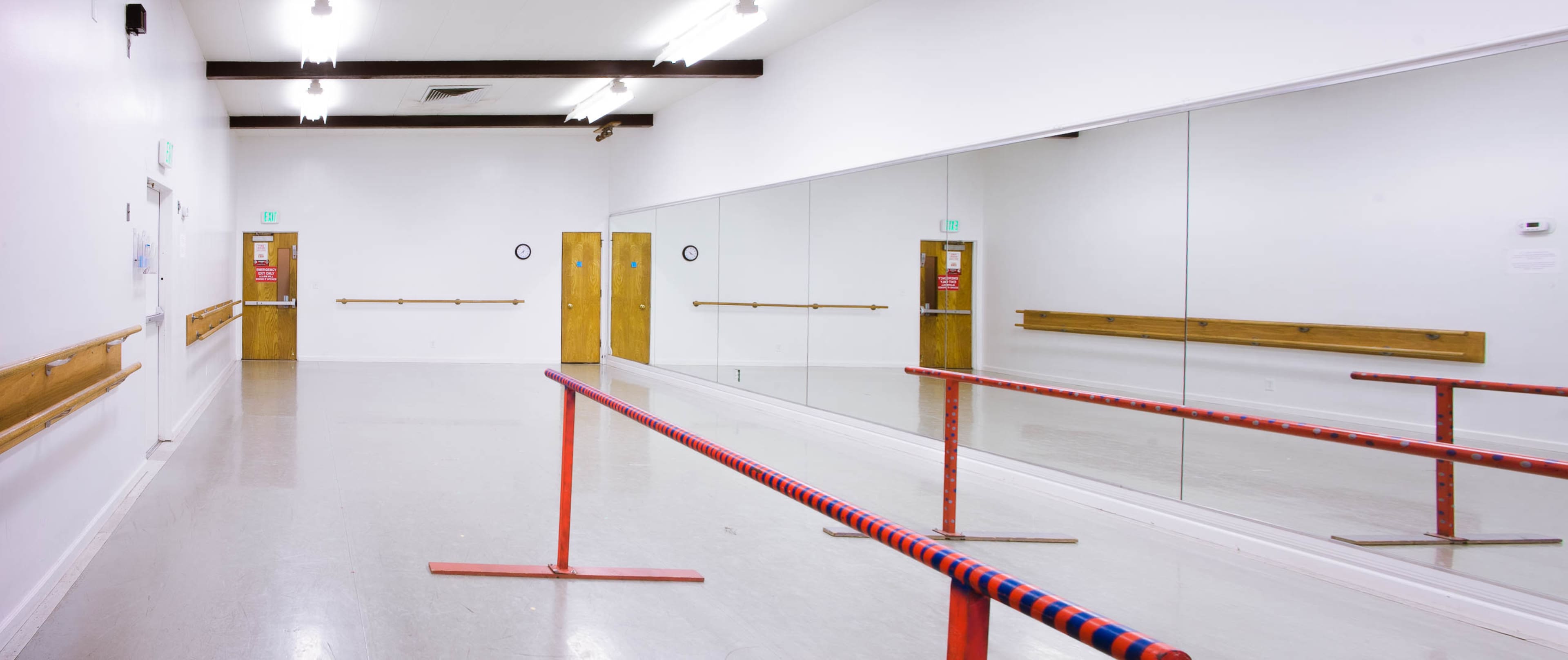 A bright dance studio features a mirrored wall, wooden barre, and double doors with exit signs.