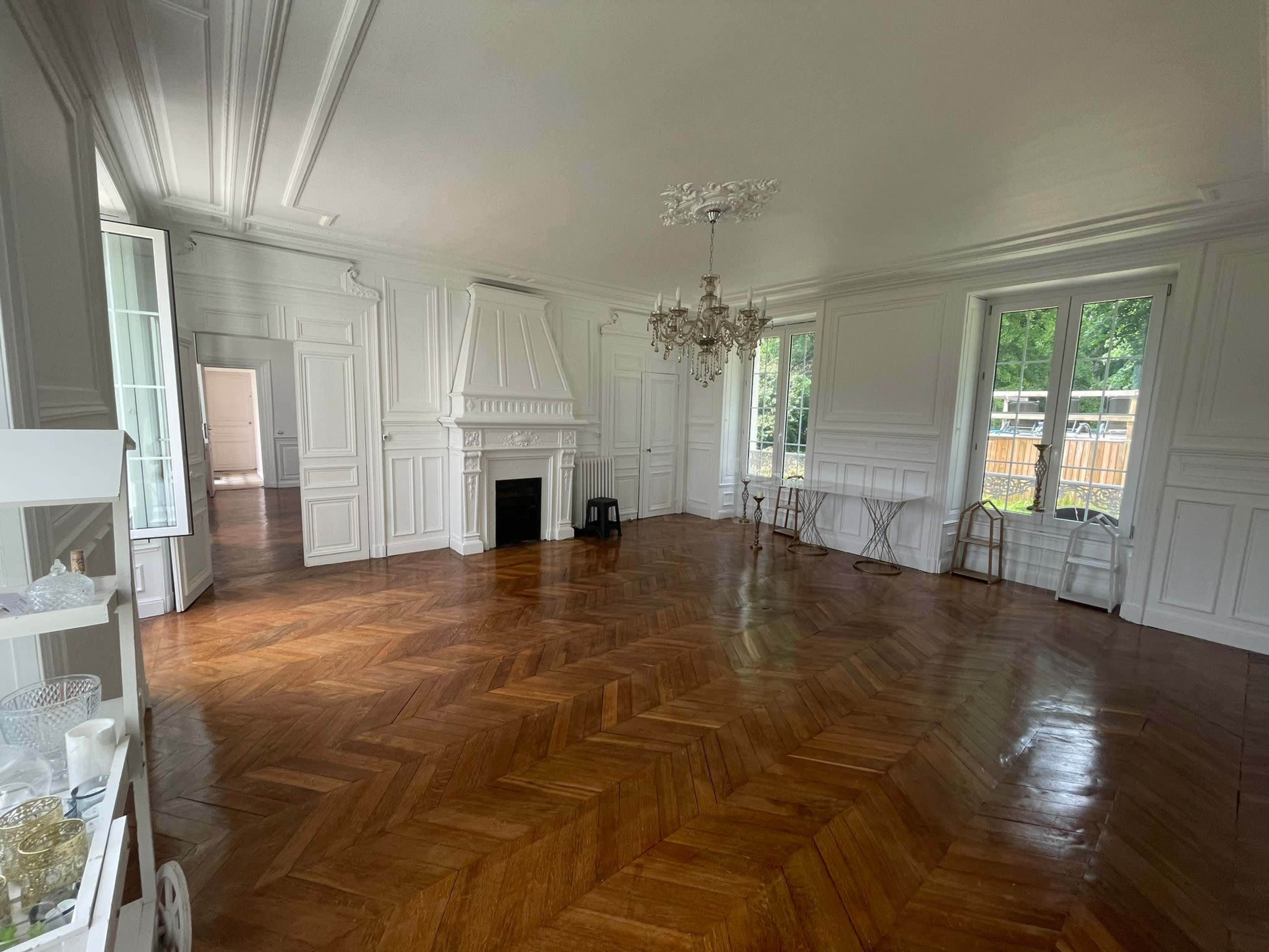 The room features a large, empty space with herringbone wood flooring, white paneled walls, a central chandelier, and a decorative fireplace.