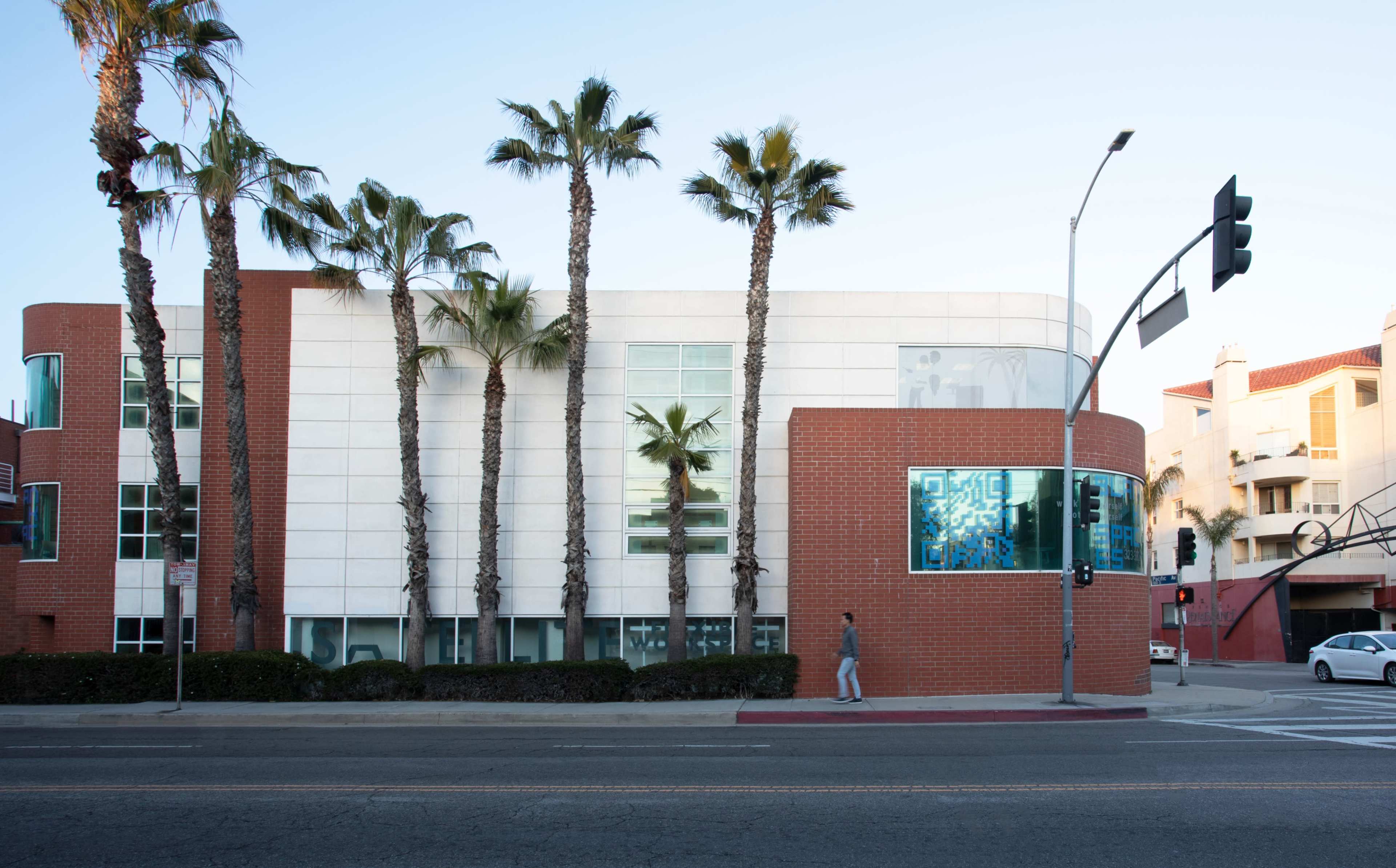 A modern building features a combination of white and brick exterior, flanked by tall palm trees along a street with a traffic signal.