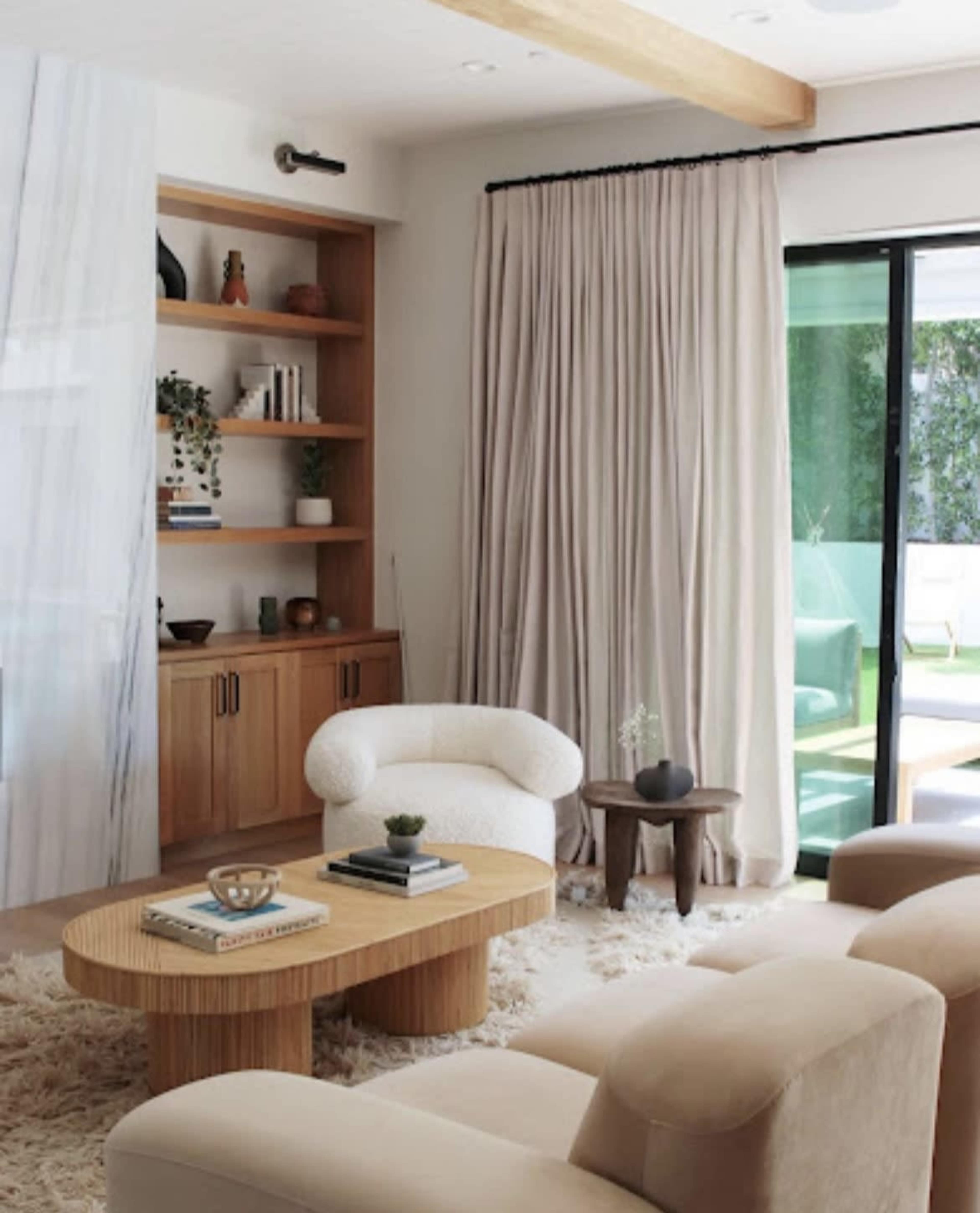 The image shows a modern living room featuring a light-colored sofa, a round wooden coffee table, a bookcase with decorative items, and large windows covered with beige curtains.
