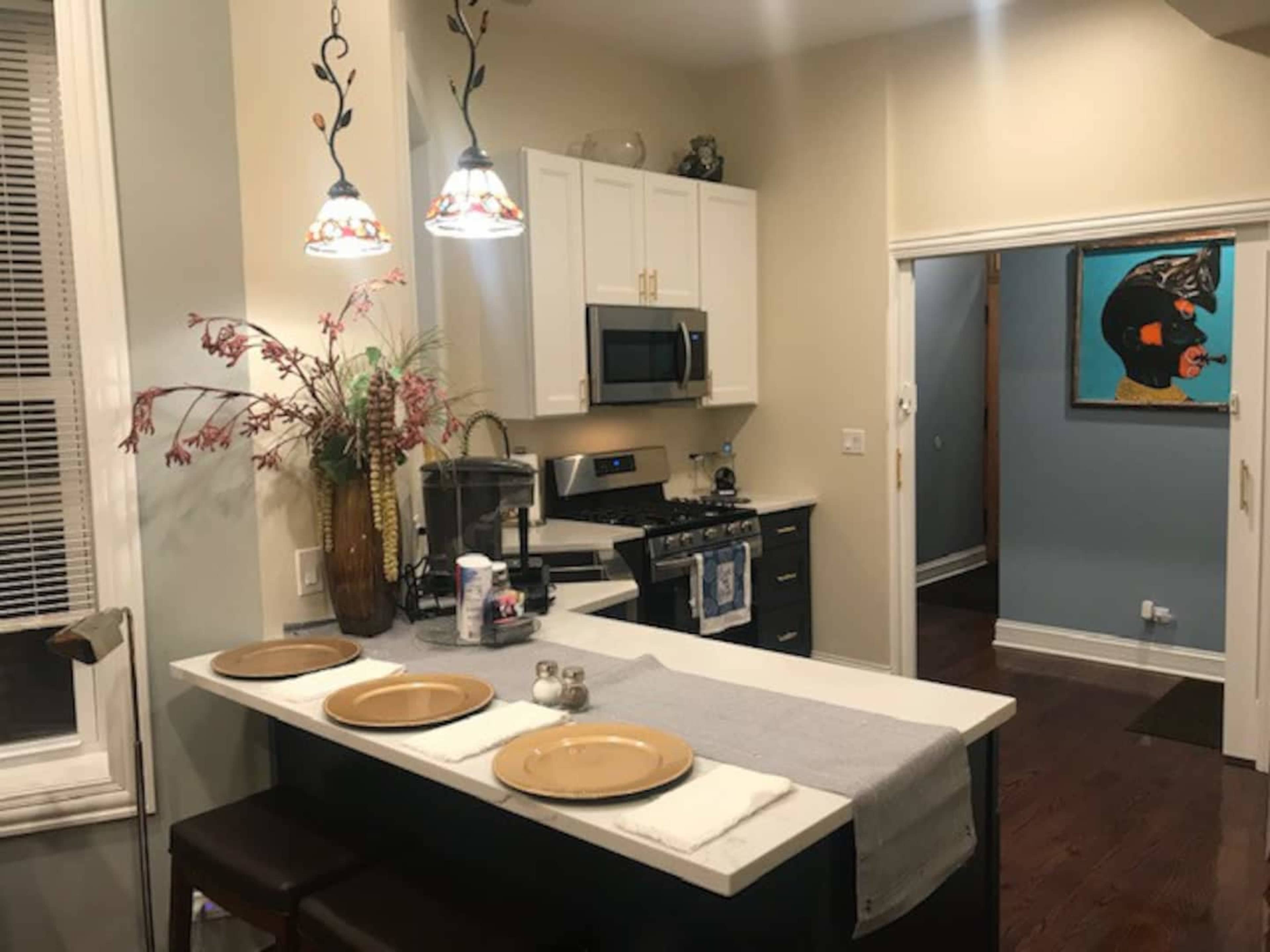 The image depicts a modern kitchen with white cabinets, a stainless steel stove, and bar seating for two, illuminated by pendant lights above the counter.