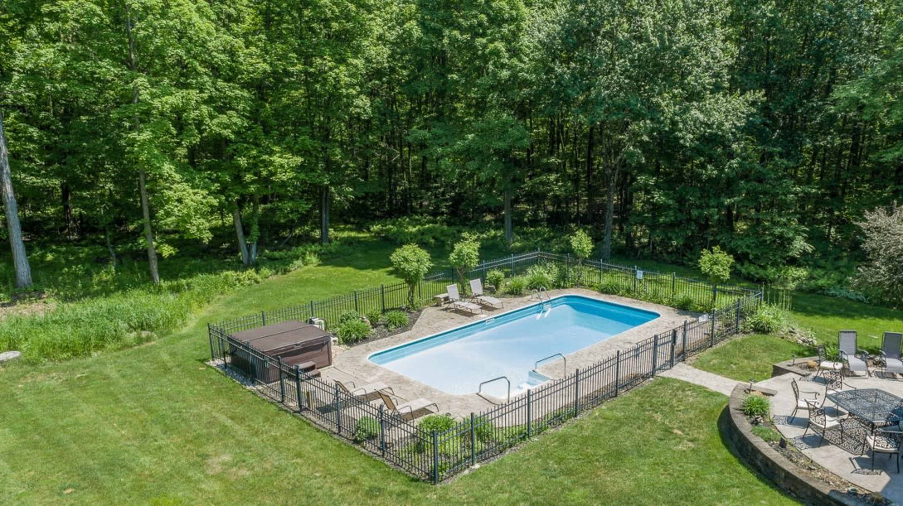 A rectangular swimming pool surrounded by a fence, with a hot tub and lounge chairs situated on a grassy area near a wooded backdrop.