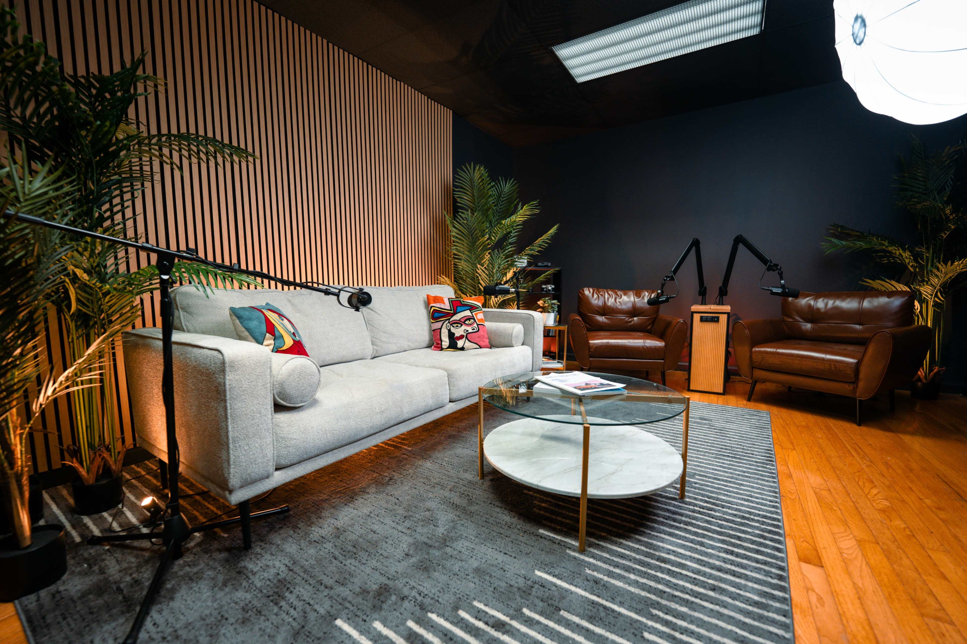 The image shows a modern podcast studio featuring a light gray sofa, two brown armchairs, a round glass coffee table, and various plants against a backdrop of wooden paneling.