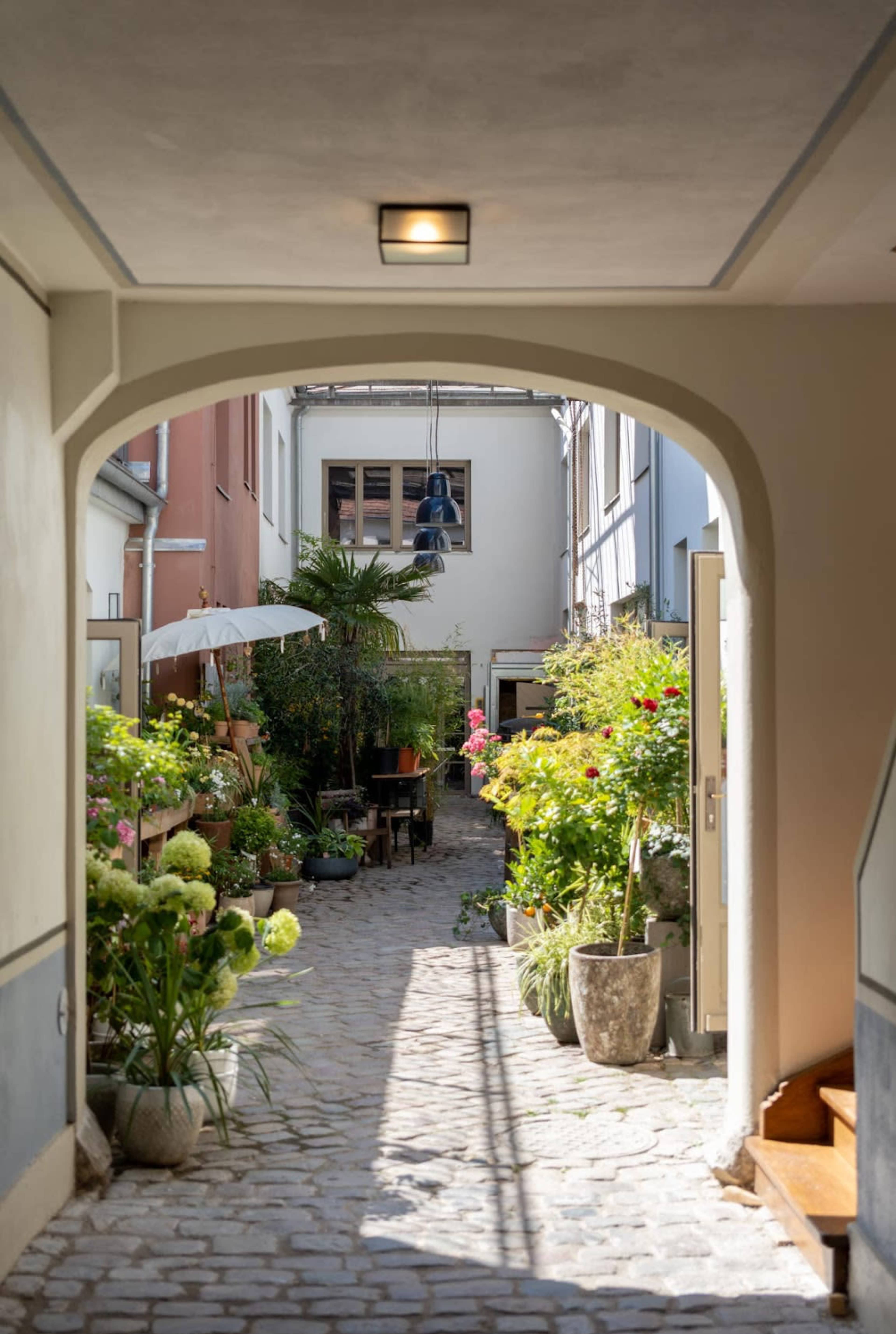 A cobblestone pathway leads through an archway into a garden courtyard filled with potted plants and flowers.