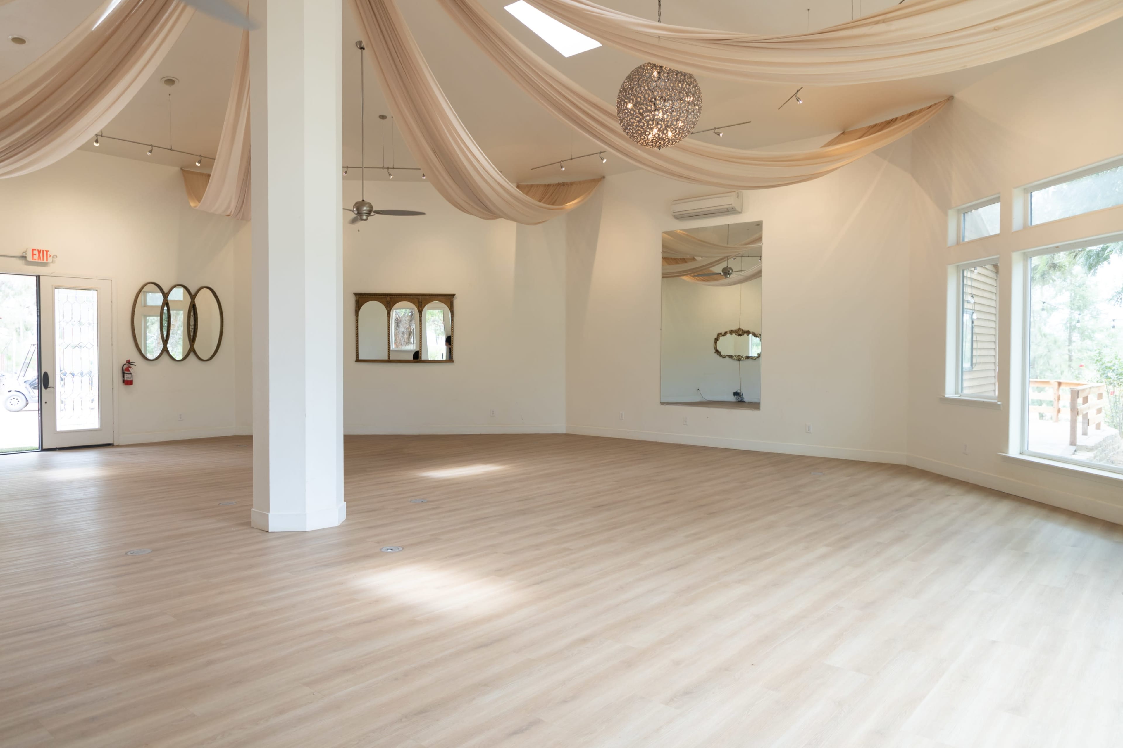 The image depicts a spacious, well-lit interior featuring wooden flooring, elegant drapery hanging from the ceiling, and large windows.
