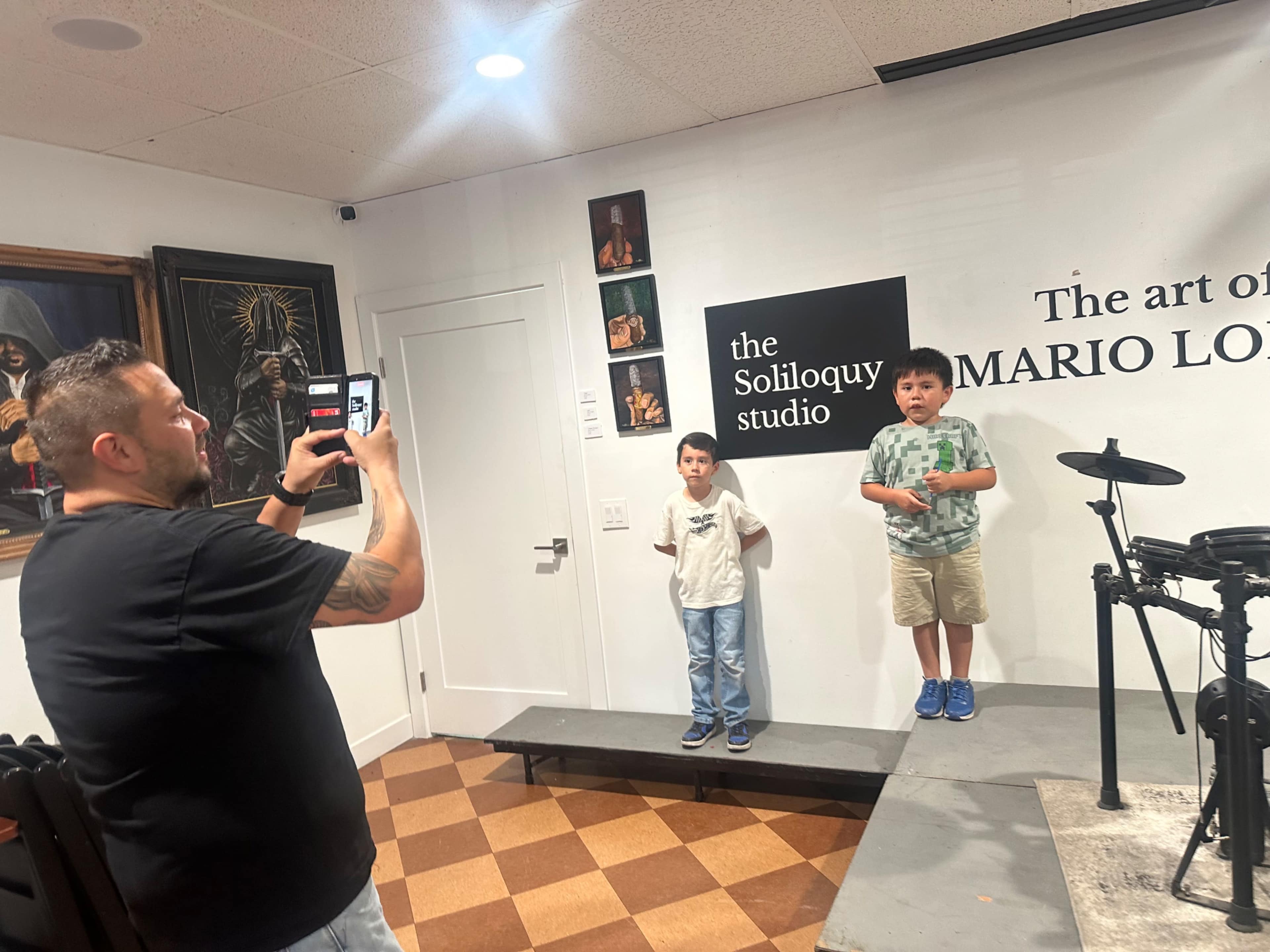 A man is taking a photo of two boys standing on a small stage in an art studio, with gallery walls displaying various artworks.