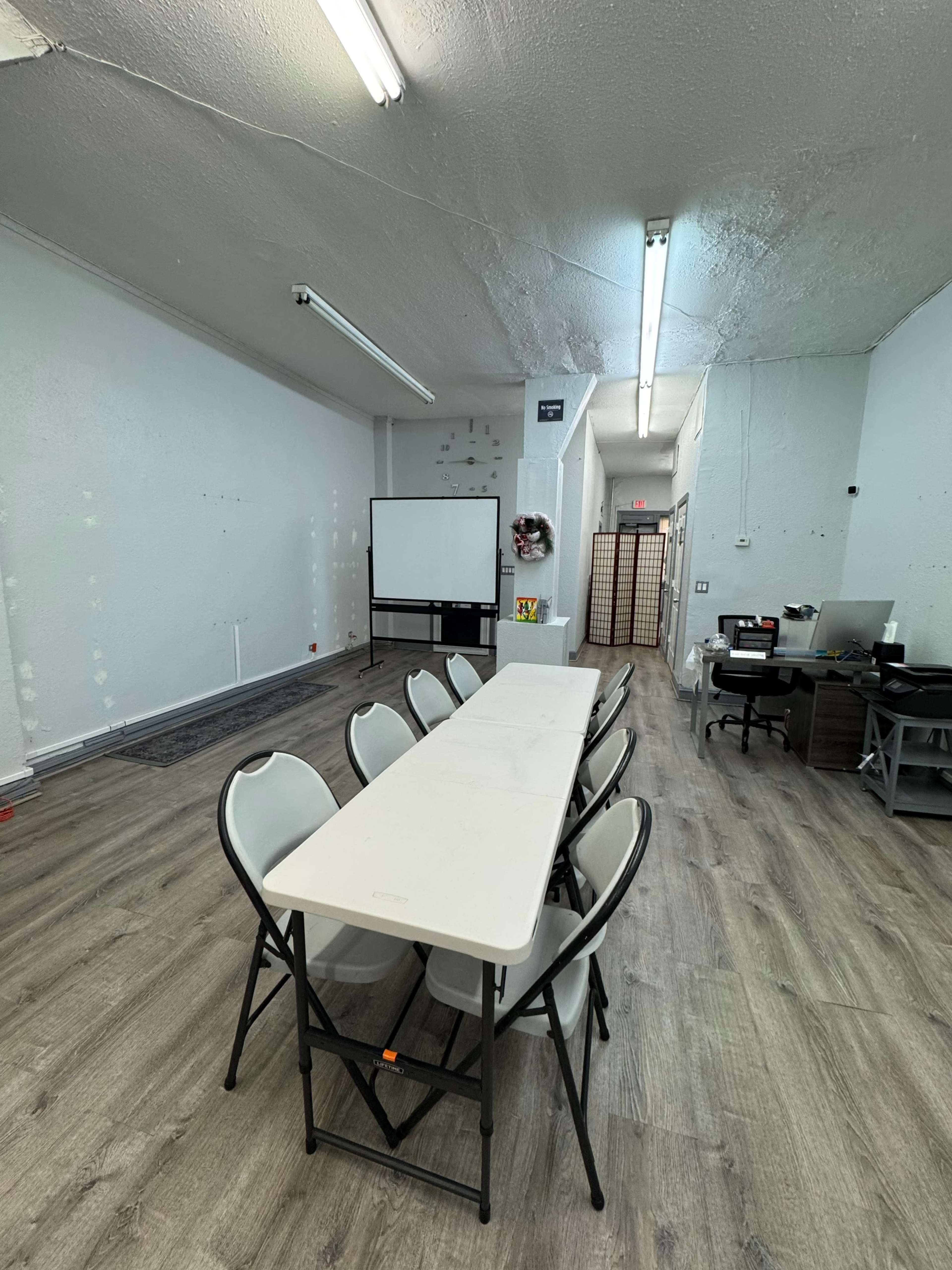 The image shows an empty room with a long table surrounded by chairs, a projector screen on one wall, and an office area in the corner.