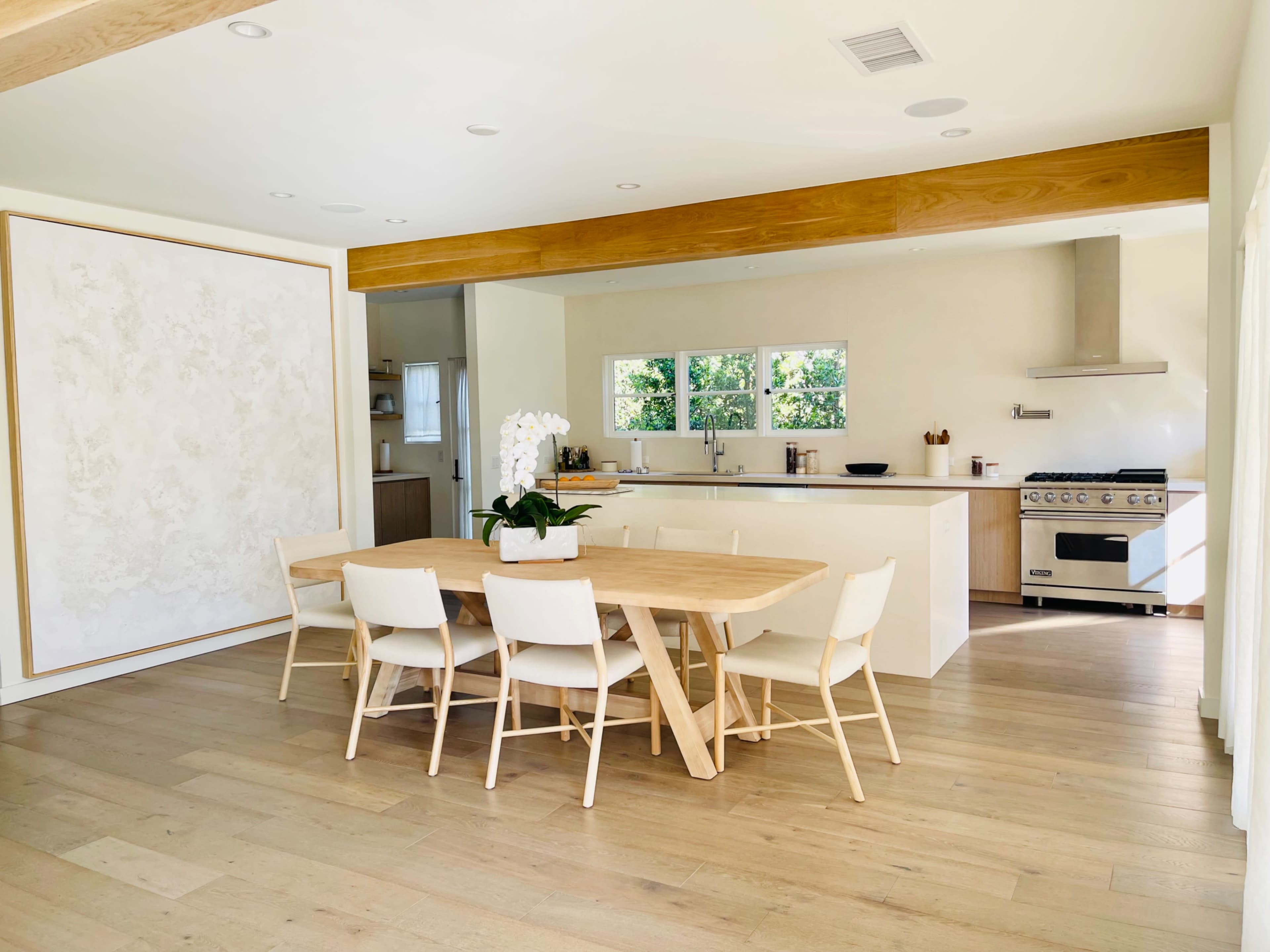 A modern kitchen and dining area feature a large wooden table surrounded by white chairs, with large windows allowing natural light and a view of greenery outside.