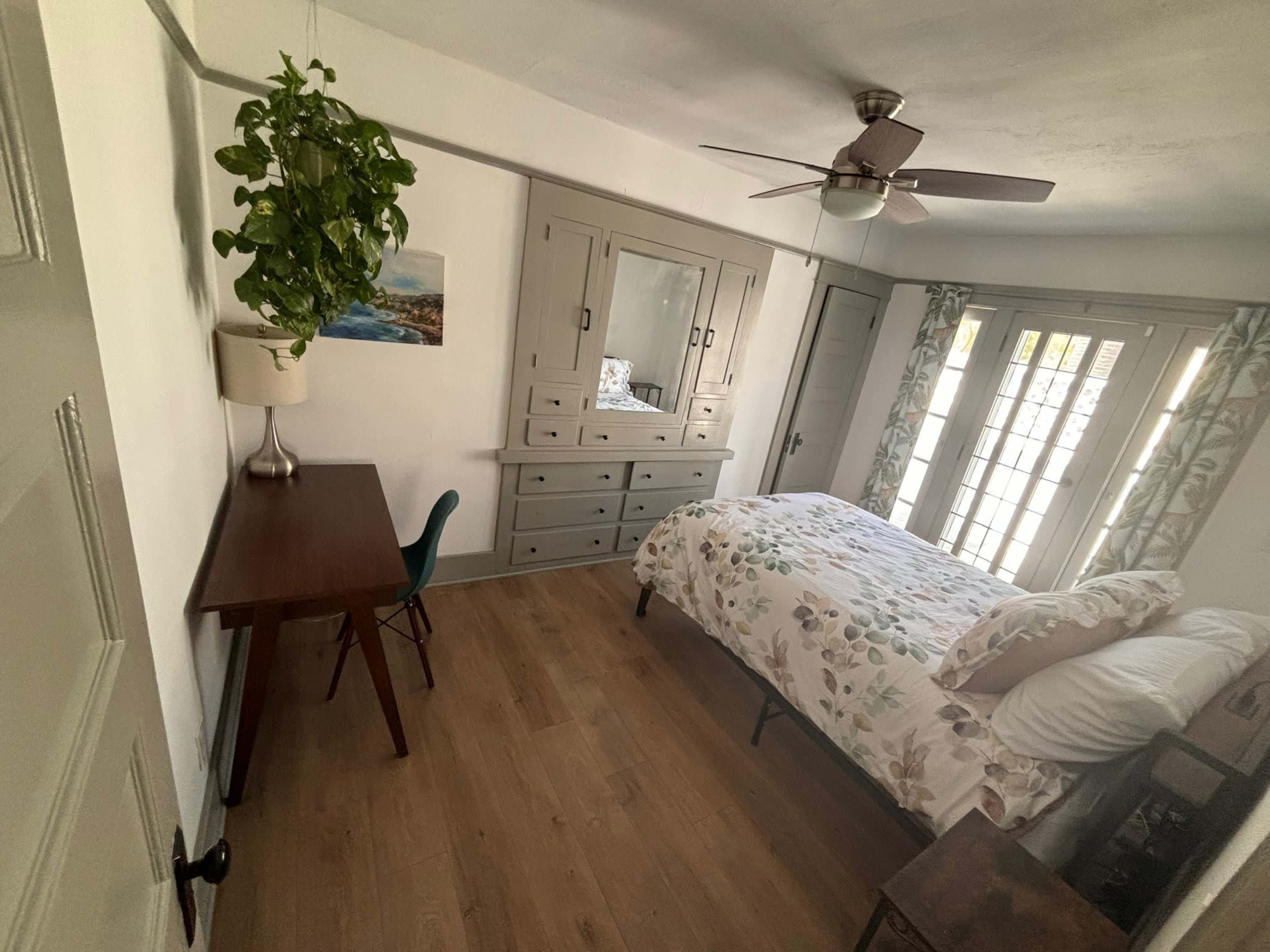 The room features a bed with a floral bedspread, a wooden desk and chair, a mirror dresser, and a plant hanging near a window with multiple panes.