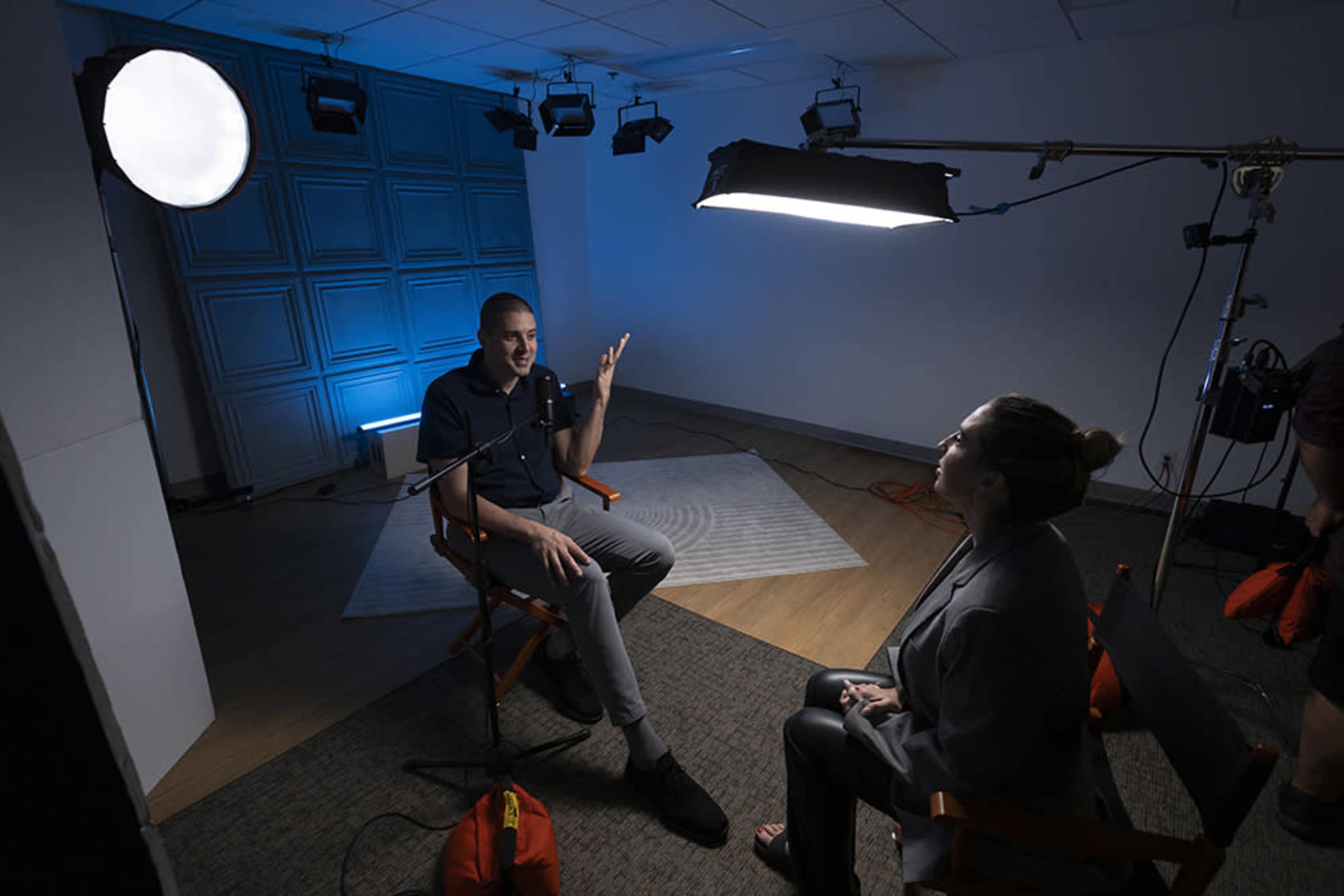 A man sits in a director's chair while speaking to a woman in a similar chair during a video interview setup with professional lighting.