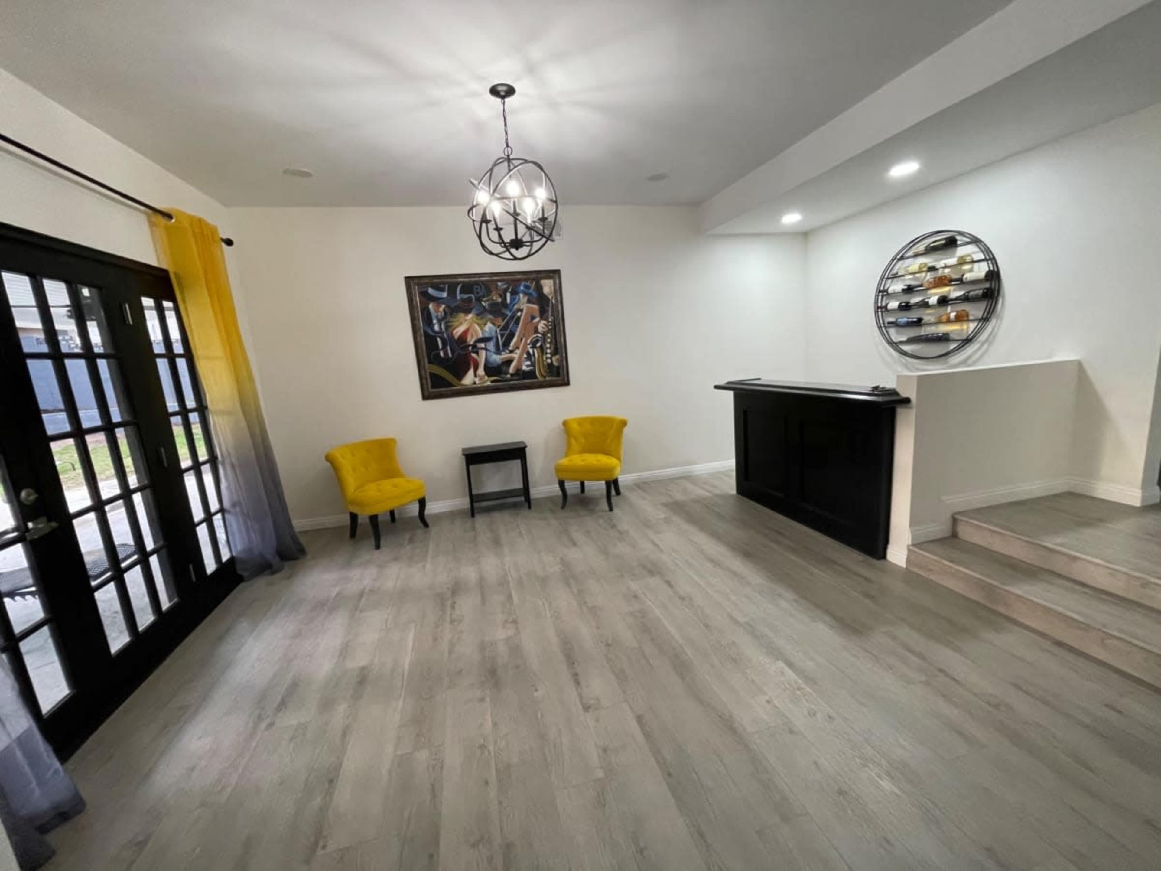 A spacious room with two yellow chairs, a small table, and a bar area, featuring a large painting on the wall and a stylish light fixture overhead.