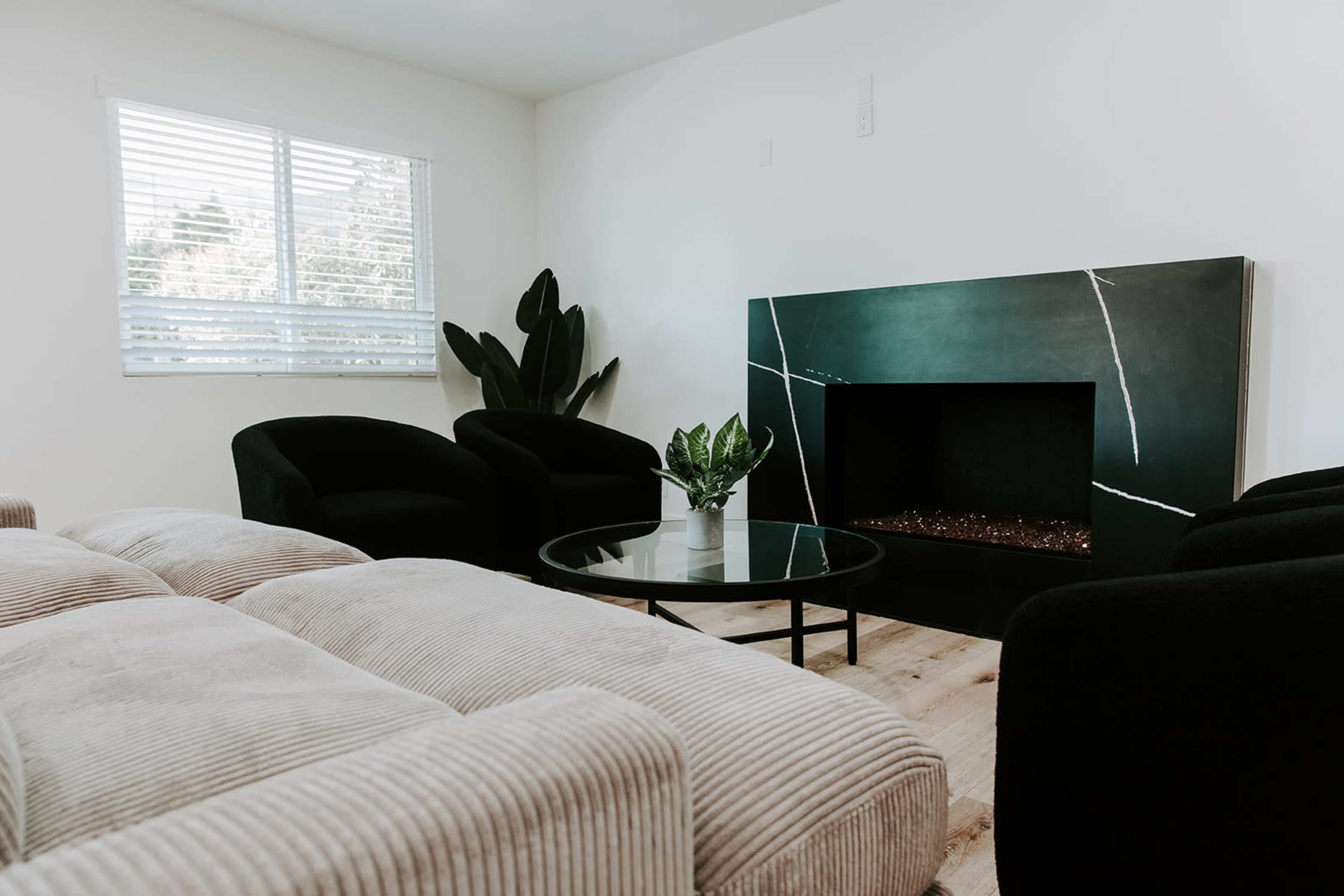 A modern living room features a black fireplace, a round glass coffee table, and three black chairs surrounded by light-colored walls and a window with blinds.