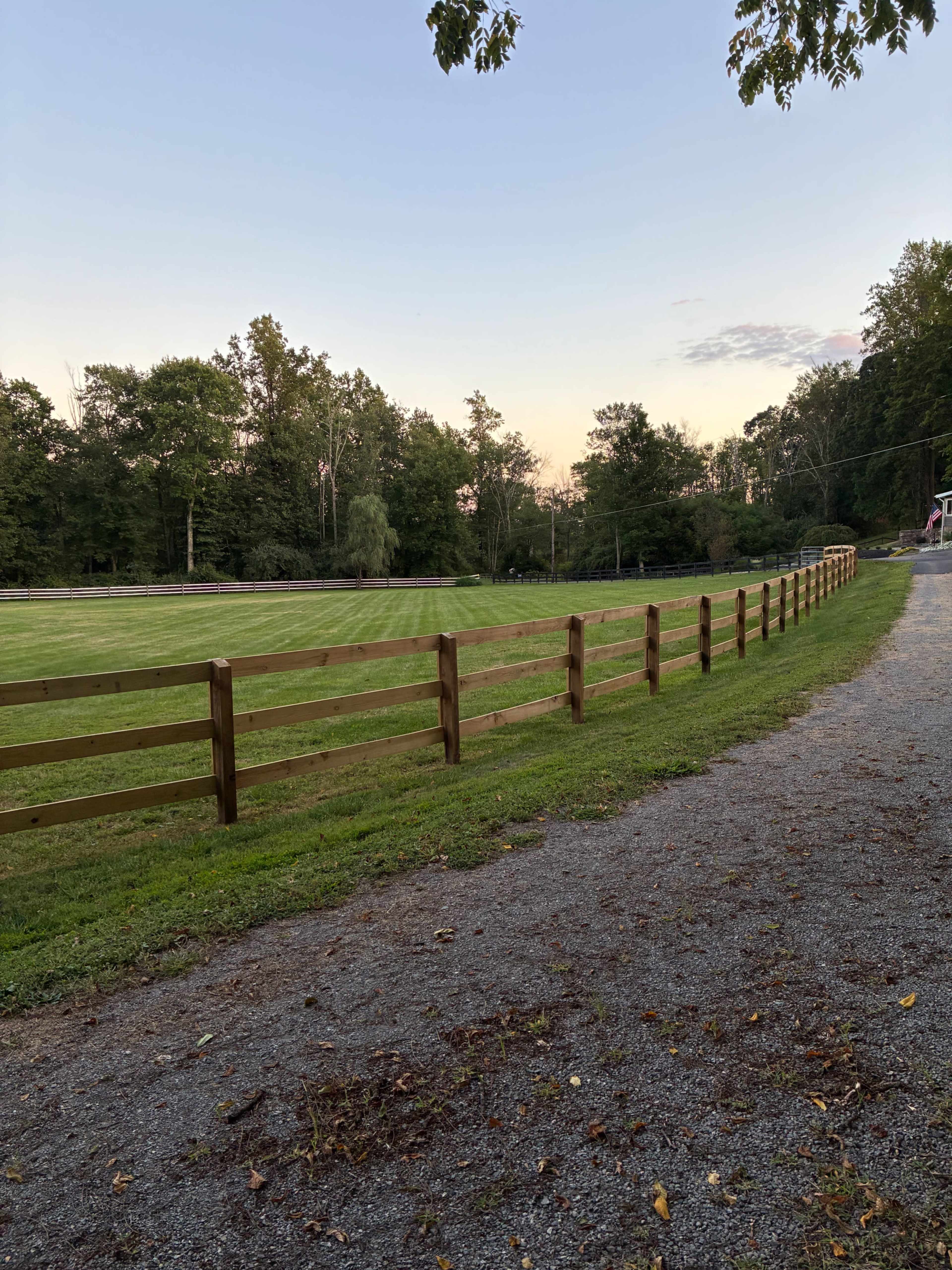 A gravel pathway runs alongside a wooden fence that borders a grassy field, surrounded by trees.