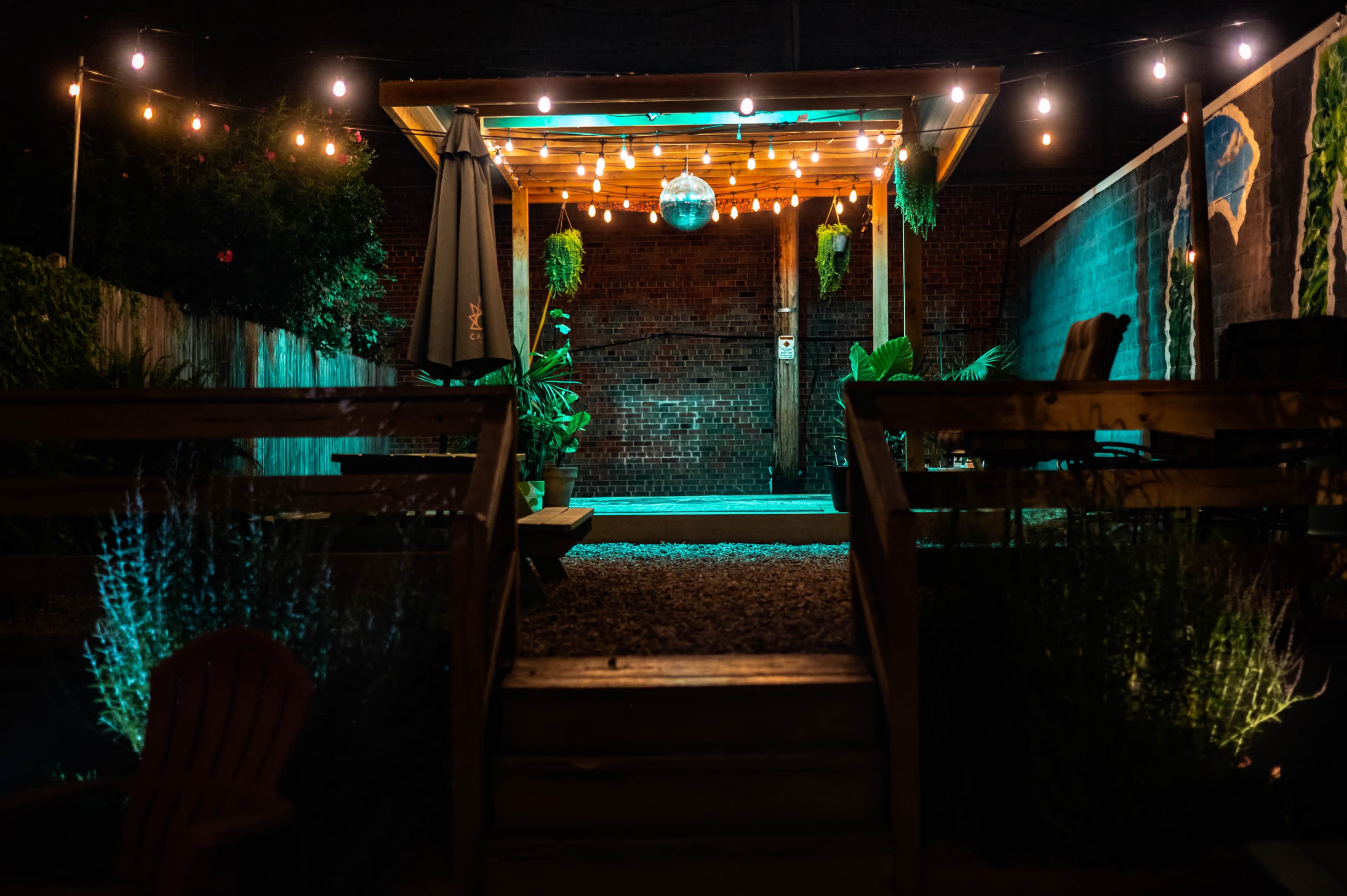A dimly lit outdoor patio area with string lights, a disco ball hanging from a wooden structure, and potted plants arranged on the sides.