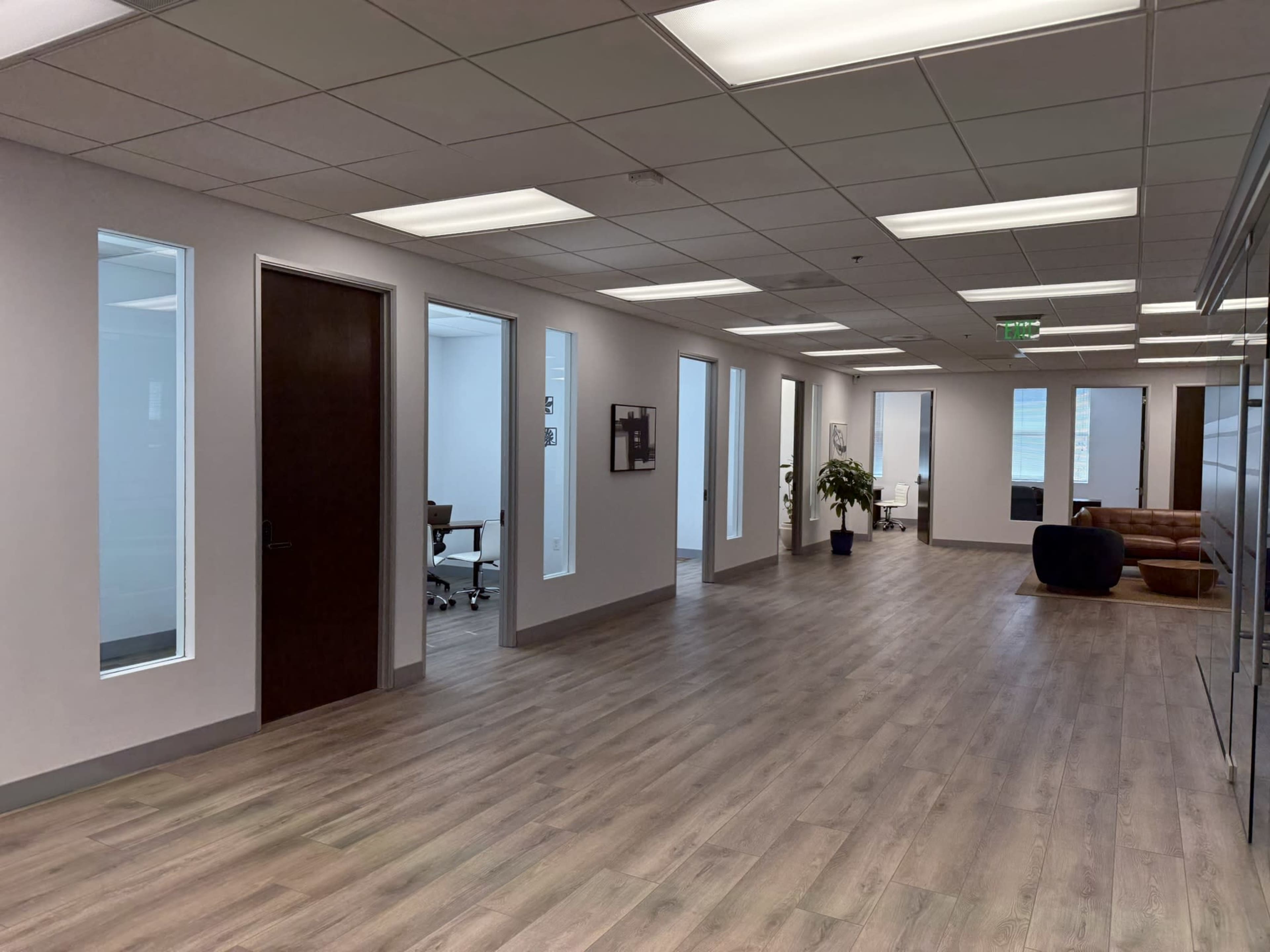 The image shows a spacious office hallway with multiple glass-fronted offices and a lounge area featuring a sofa.