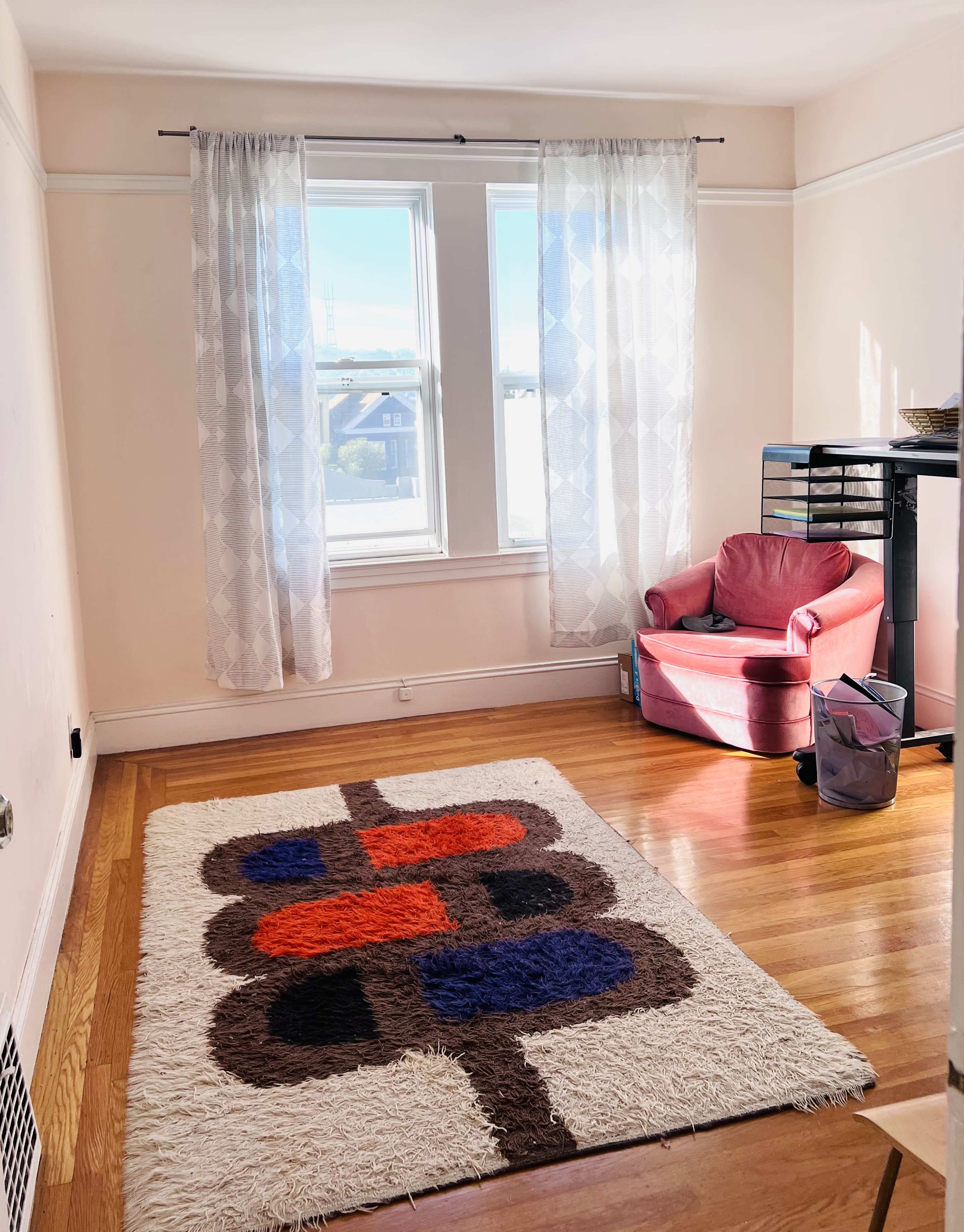 A bright room features a large window, a patterned area rug on the wooden floor, and a pink chair next to a small wastebasket.