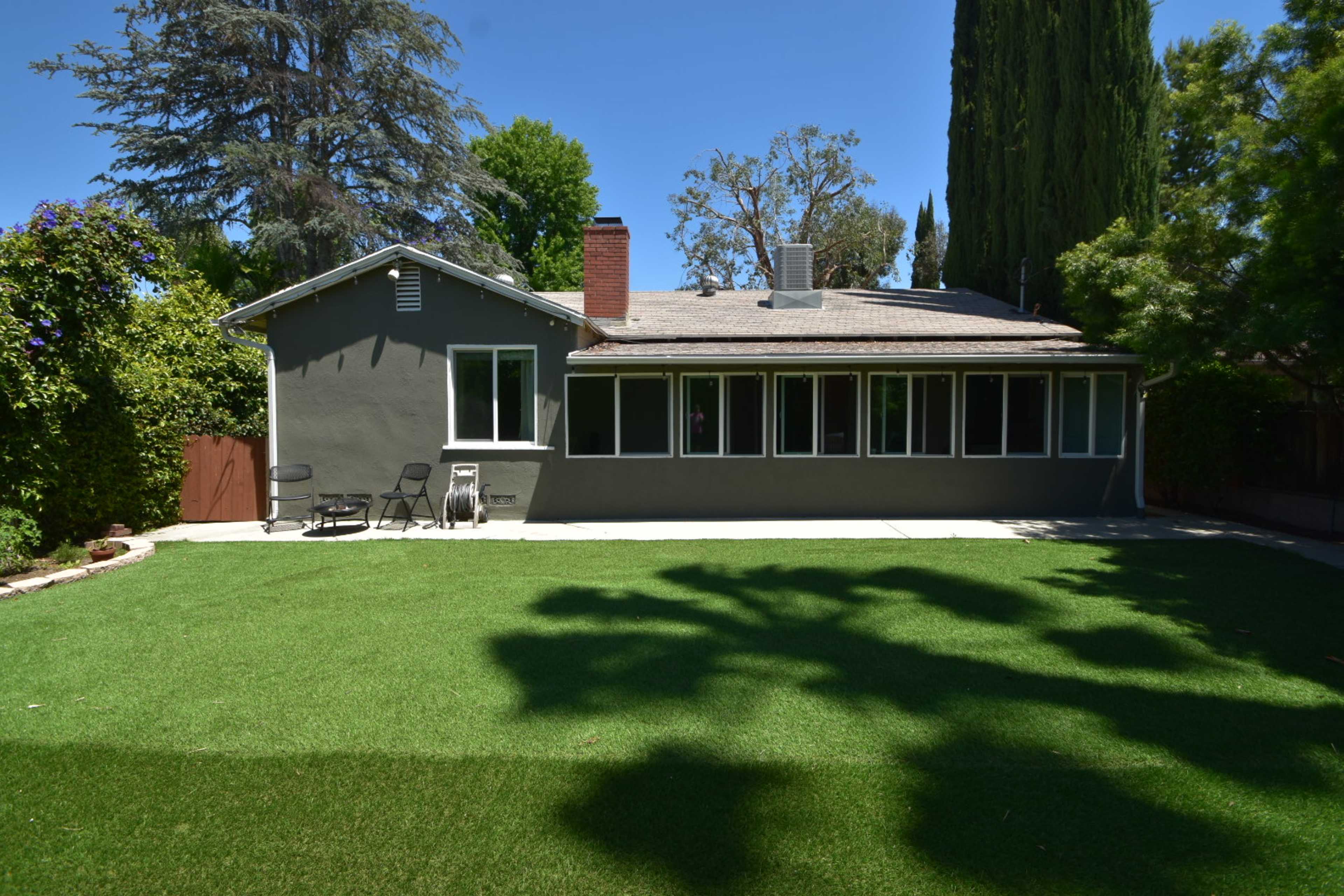 A gray house with multiple windows sits in a backyard featuring artificial grass and trees.