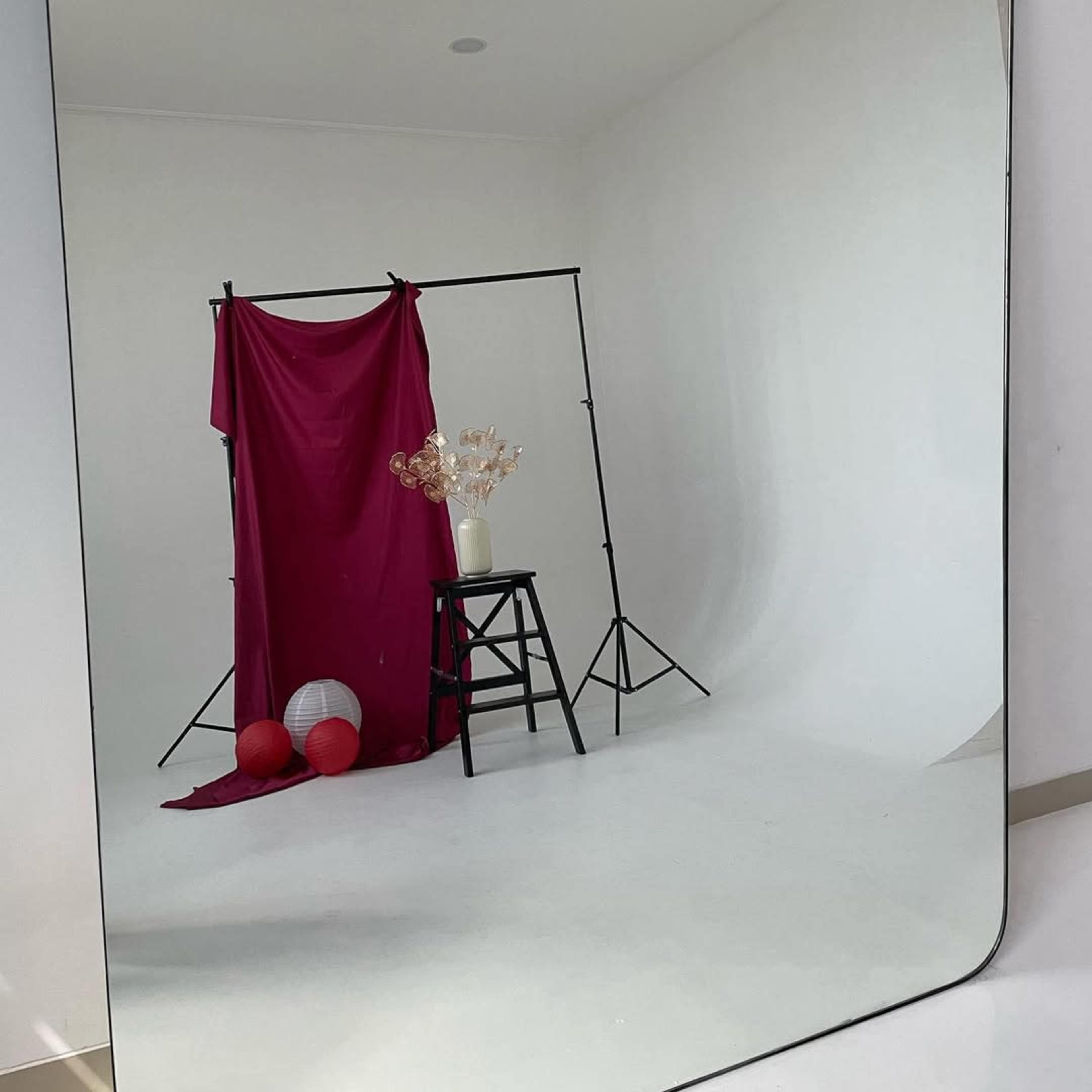 The image shows a minimalist studio setup featuring a maroon backdrop draped on a support frame, a black stool, decorative flowers in a vase, and red and white spherical props on a white floor, all reflected in a large mirror.