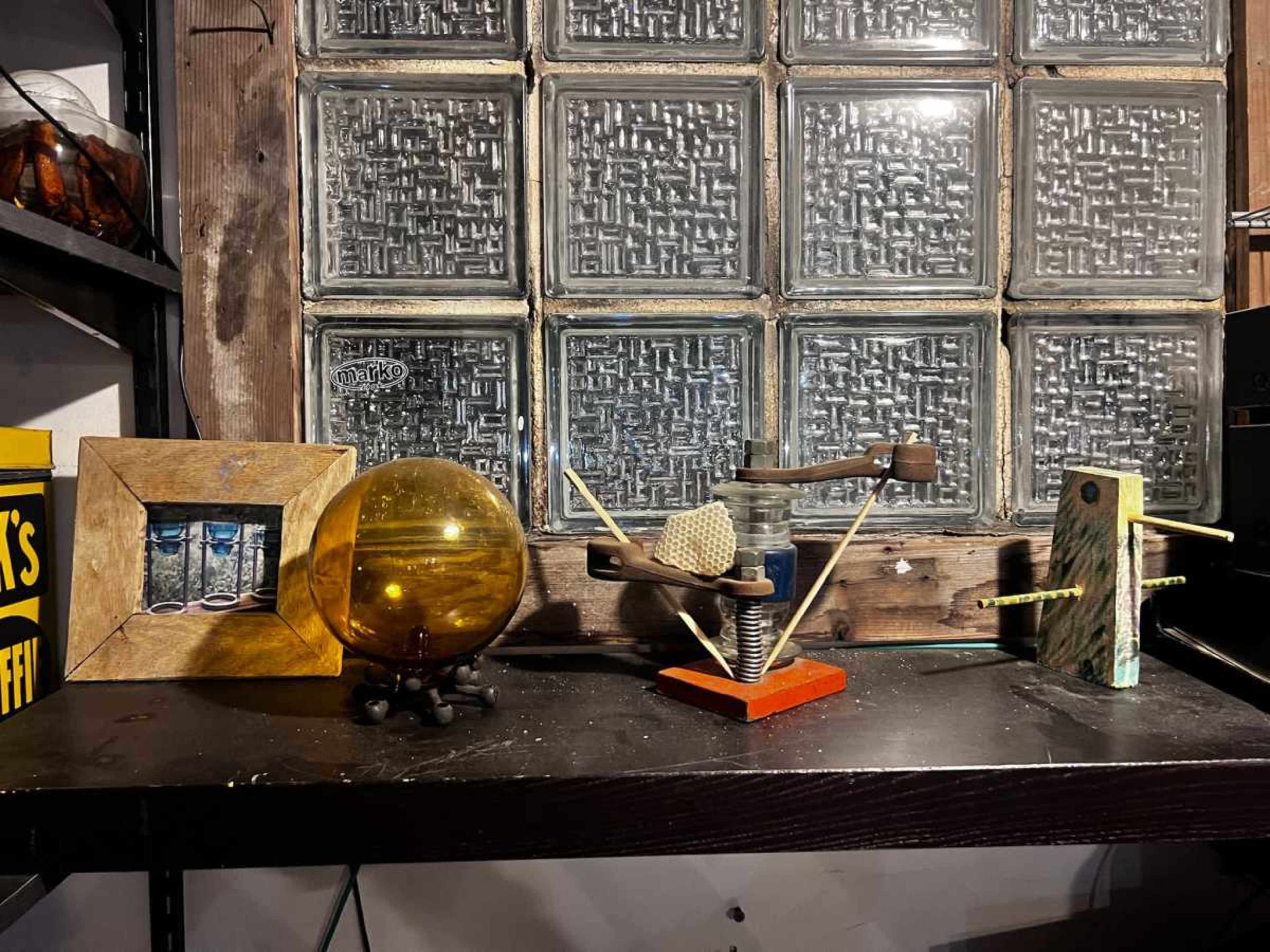 The image shows a wooden shelf displaying various objects, including a framed picture, a round glass orb, a mechanical device, and a wooden block with protruding elements, all against a backdrop of a textured glass window.