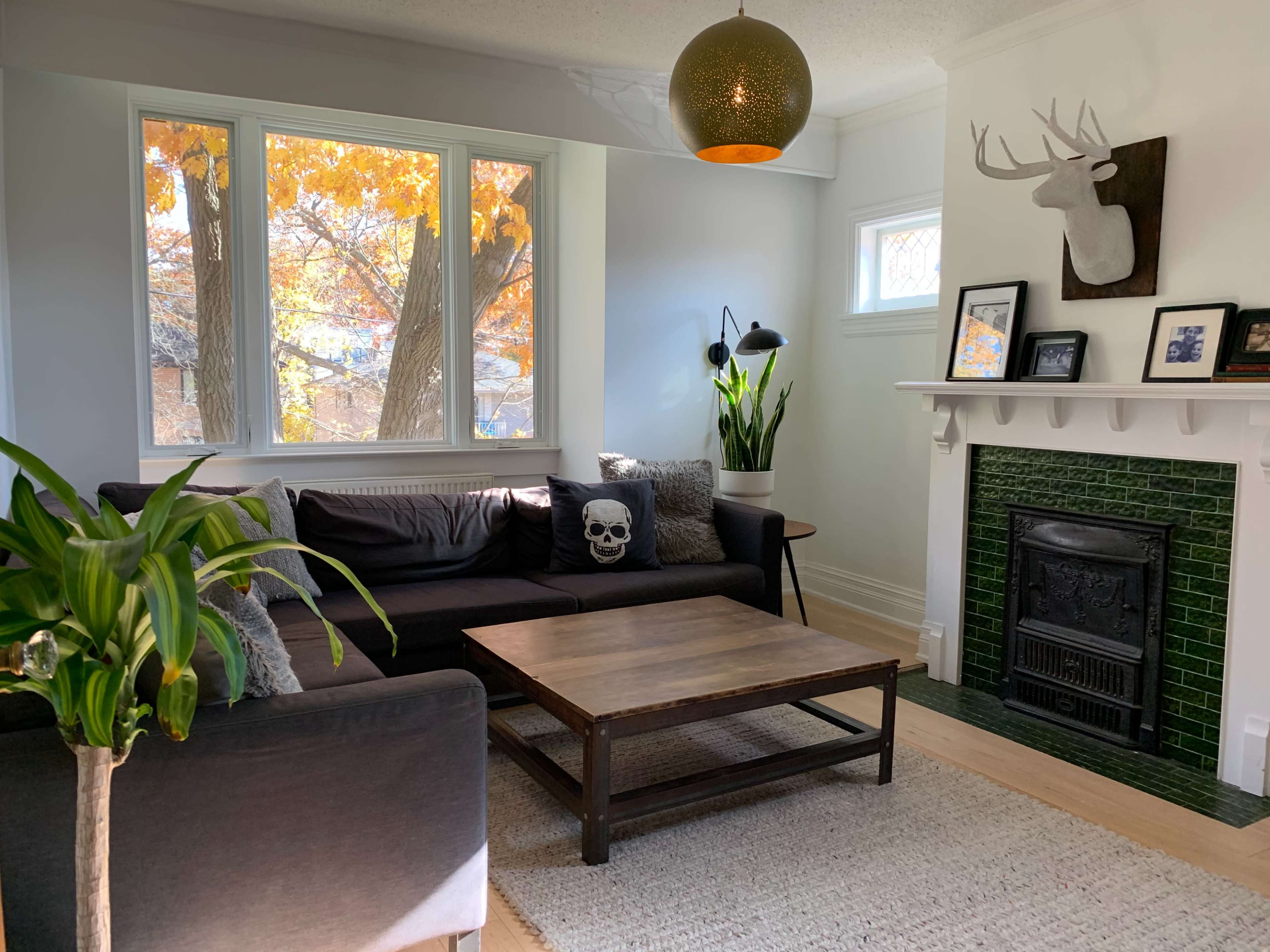A cozy living room features a dark sofa with a skull pillow, a wooden coffee table, plants, and a fireplace with green tiles, all set against large windows showing autumn trees outside.