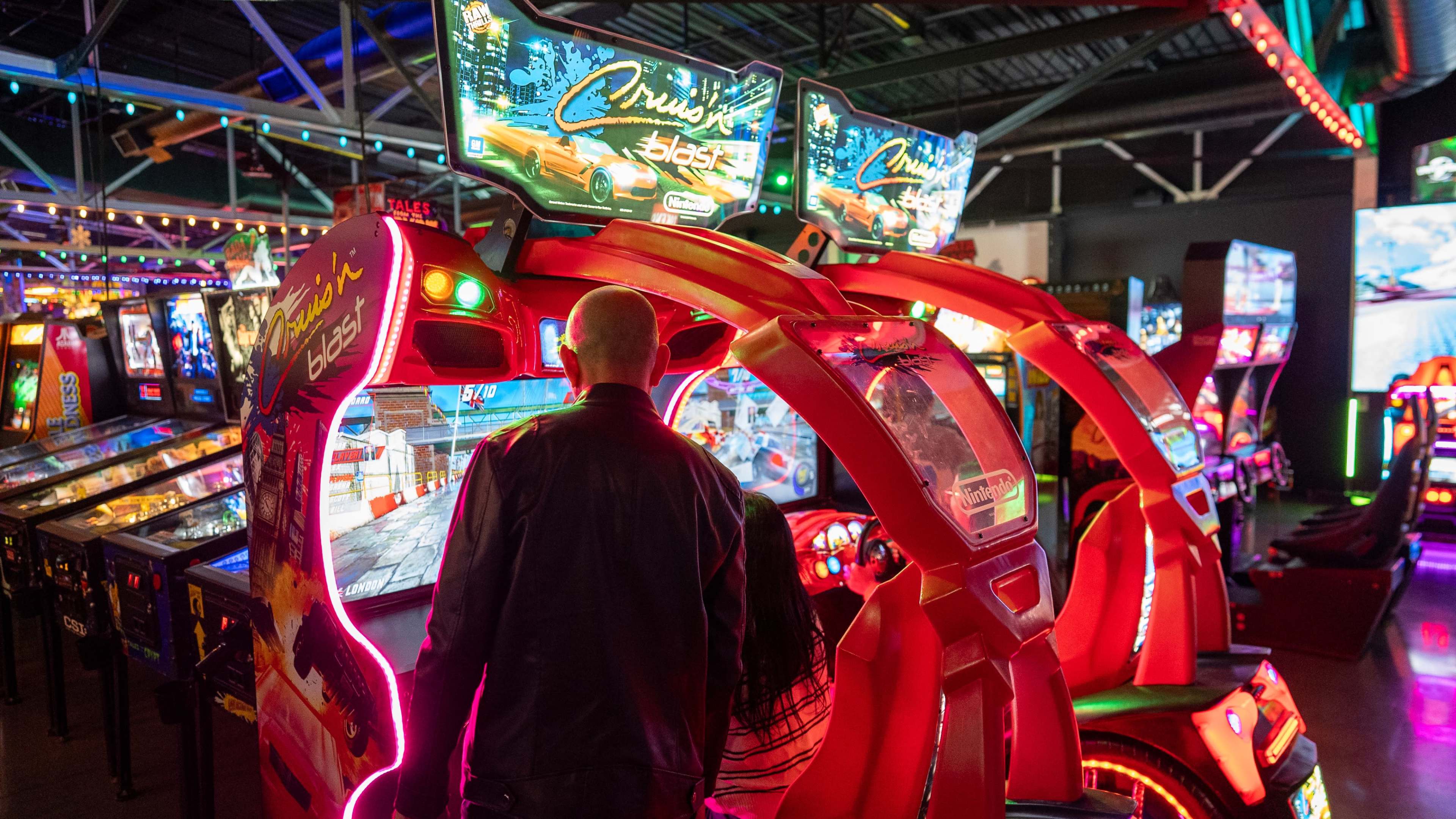 A person and a child play a racing arcade game surrounded by colorful lights and other arcade machines.